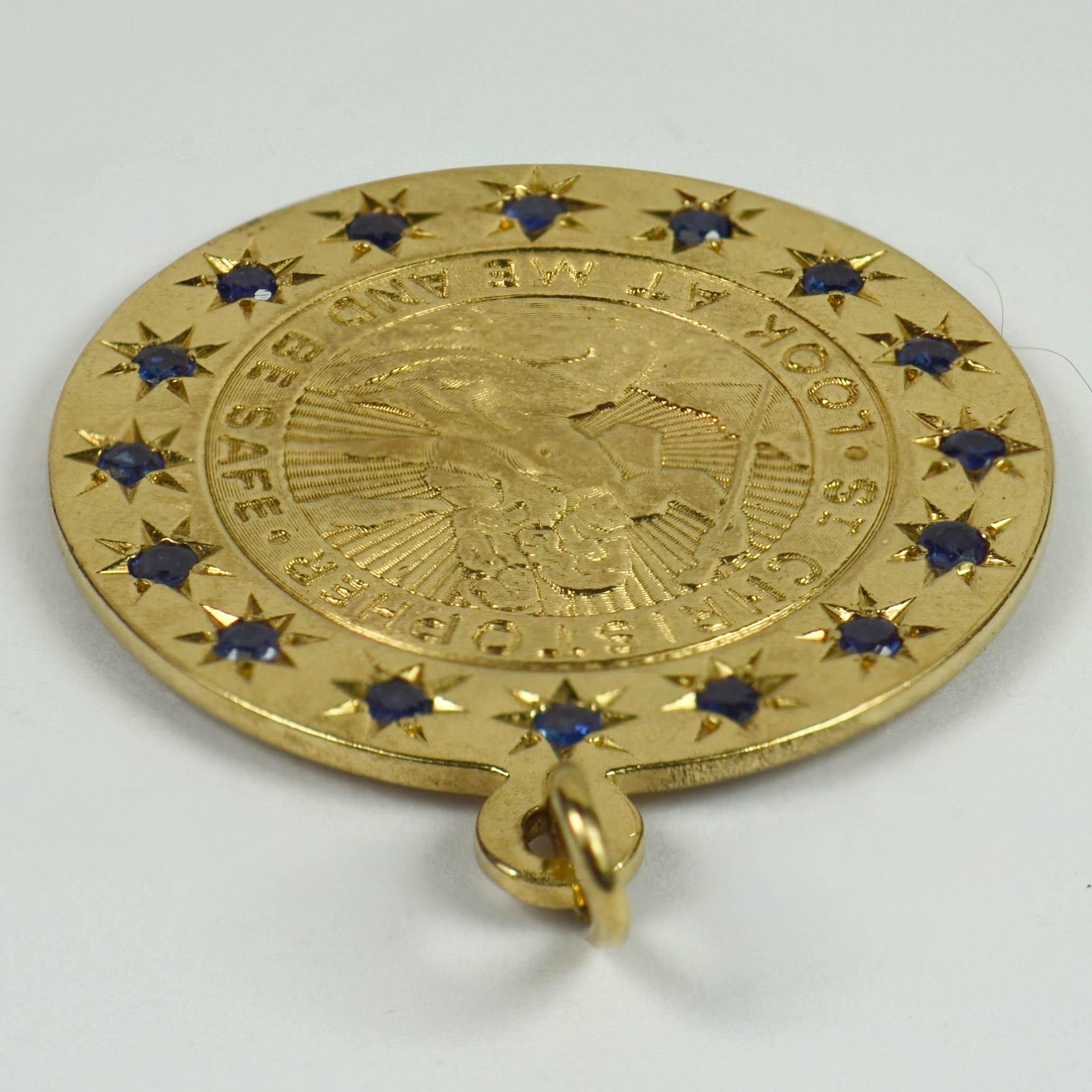 A 14 karat yellow gold charm pendant designed as a medallion depicting St Christopher carrying the infant Christ across a river surrounded by 16 blue sapphires in star settings. The pendant is engraved with the phrase 