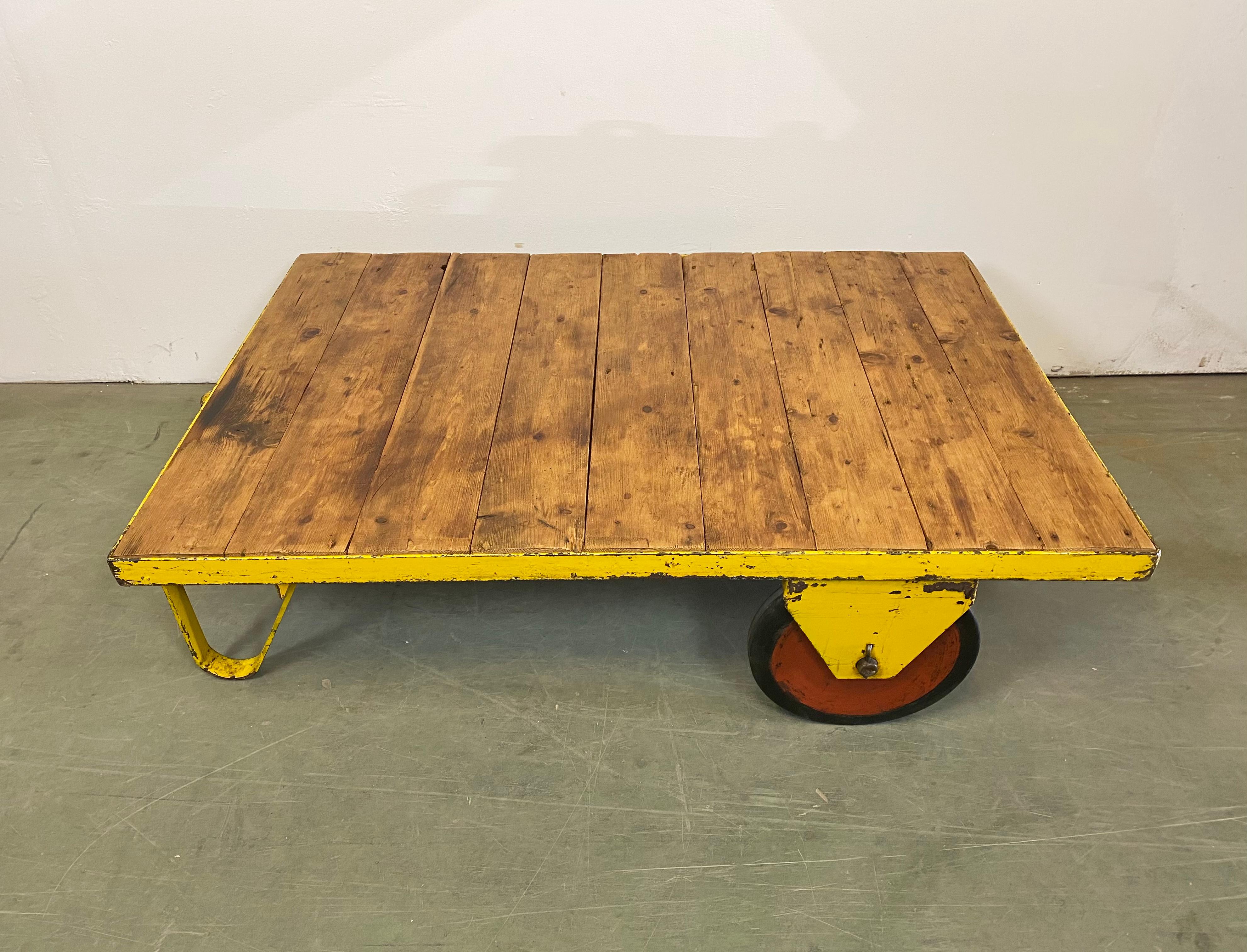 Former pallet truck from a factory now serves as a coffee table. It features a yellow iron construction with two original wheels and a solid wooden plate. The weight of the table is 41 kg.
