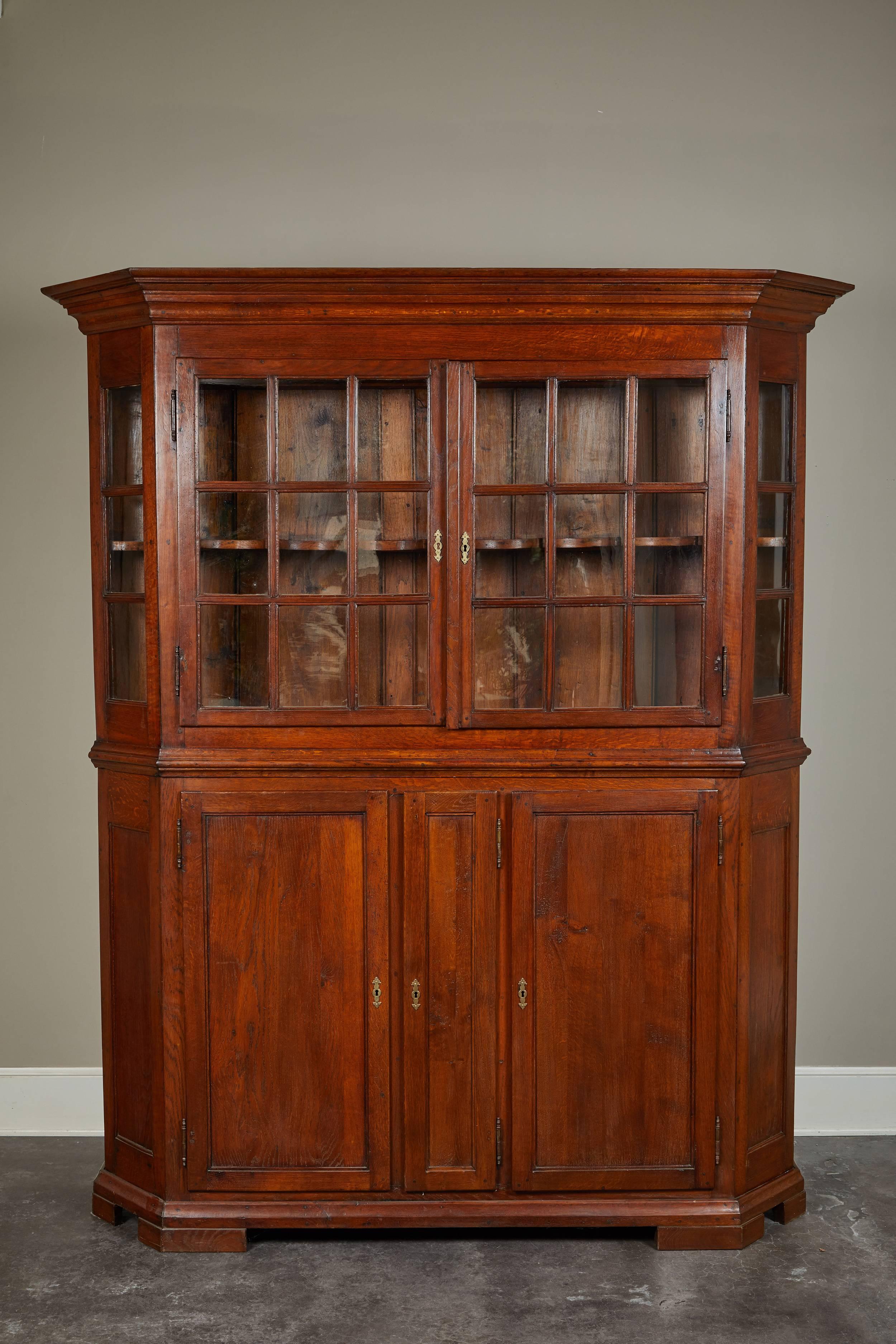 Danish baroque display cabinet, original glass and hardware, molded crown, top section consists of a pair of six paneled glass doors and two canted glass side panels enclosing one stationary centered shelf with scalloped edge and one “spoon shelf”.