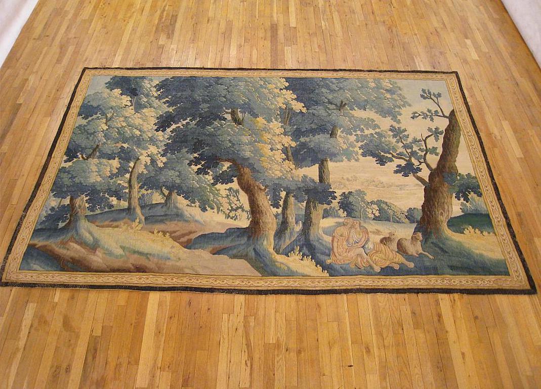 A Flemish pastoral landscape tapestry from the late 17th century, depicting a shepherd and shepherdess reposing in the shade with their sheep at right, within a tranquil wooded landscape, with rolling hills in the distance. Enclosed within a