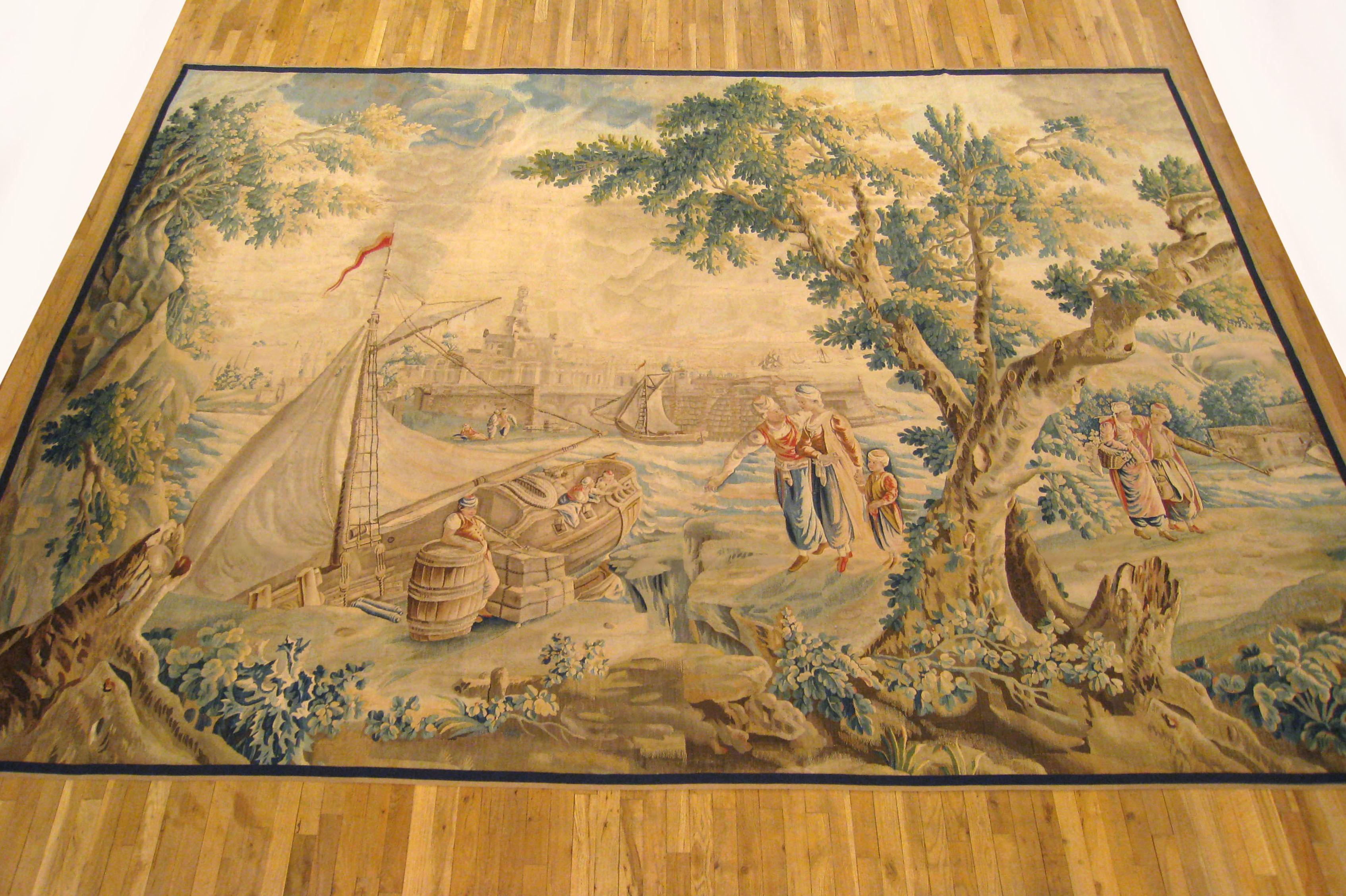 A French Aubusson rustic tapestry from the late 17th century, depicting several villagers on either side of a large tree in the right foreground, watching the cargo being unloaded from a large vessel docked by the verdant river bank at left, with