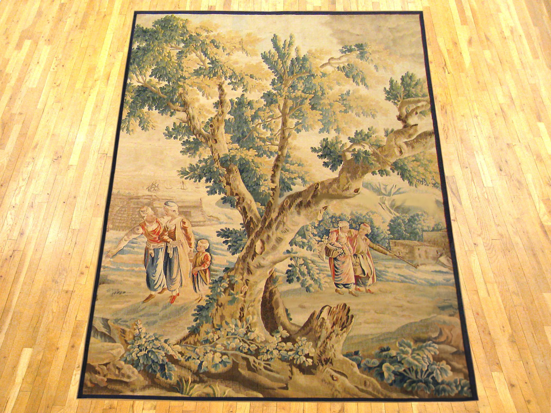 A French Aubusson rustic tapestry from the late 17th century, depicting several villagers on either side of a large tree in the right foreground, watching the cargo being unloaded from a large vessel docked by the verdant river bank at left.