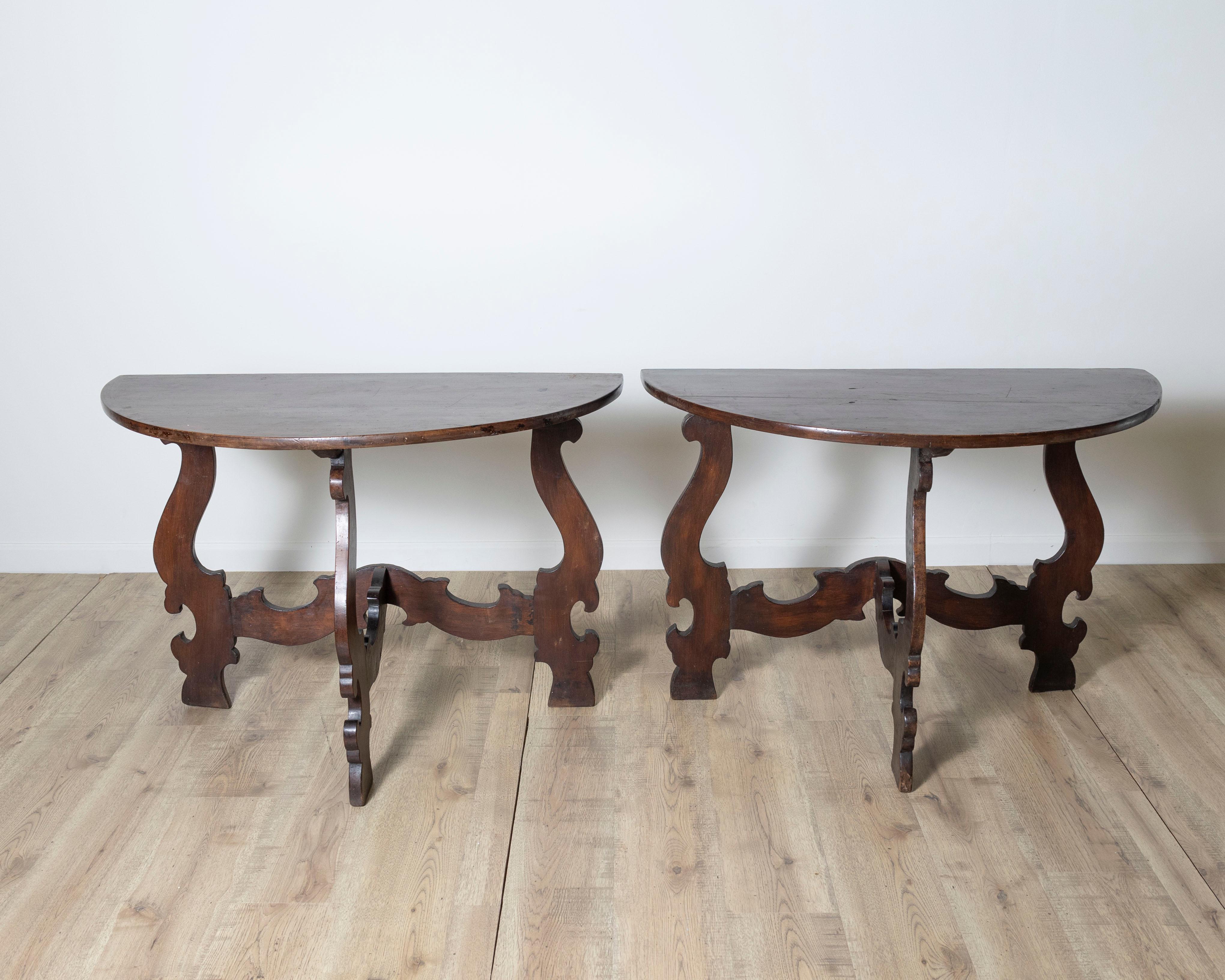 An exceptional pair of hand-carved console tables in solid walnut, crafted in Tuscany in the late 17th century. These elegant consoles feature refined semicircular tops supported by boldly sculpted lyre-shaped legs, joined by a central stretcher