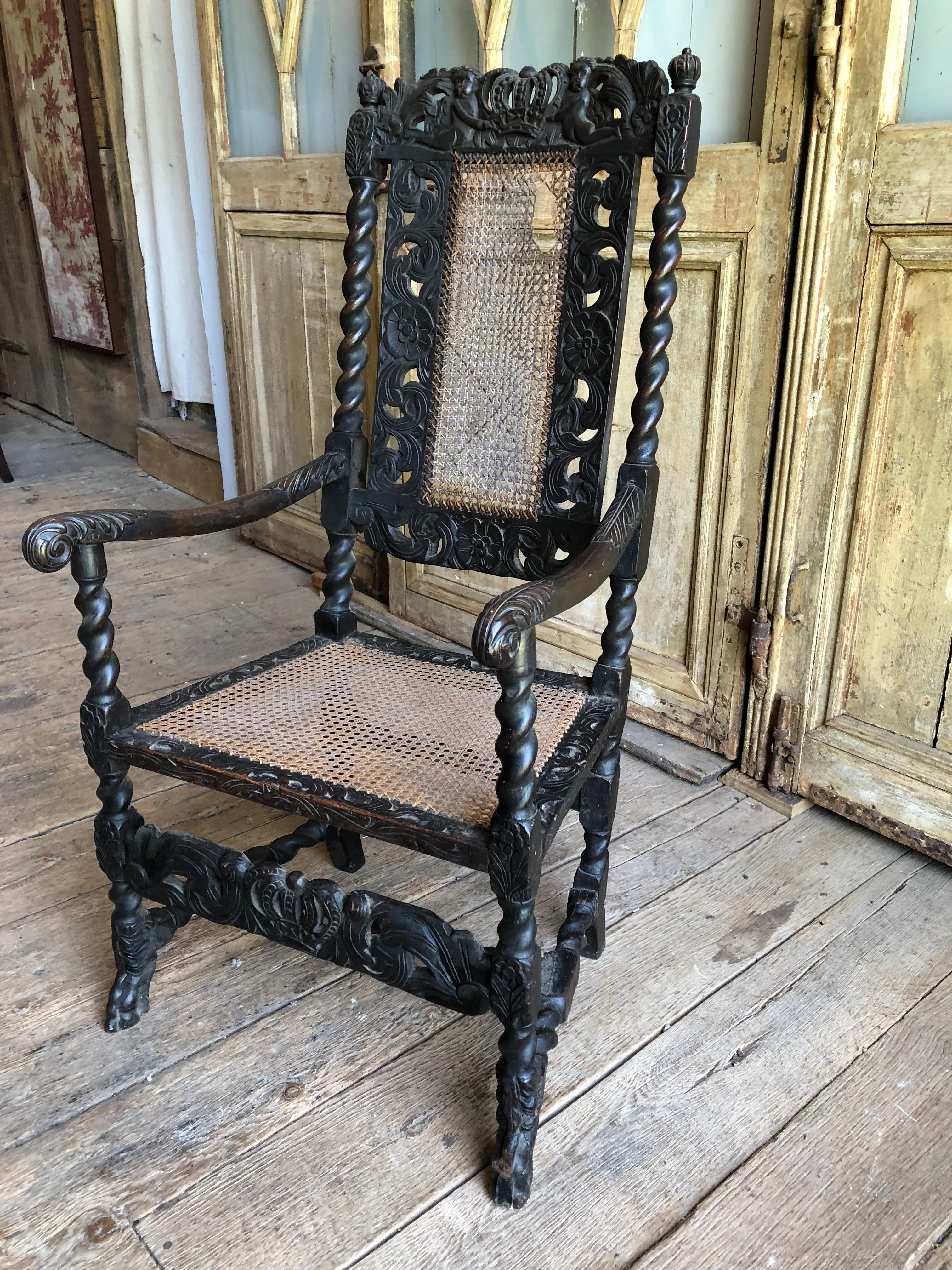 An English, William and Mary Period armchair with caned seat and back, circa 1690-1720, with elaborately carved crest rail with cherubs holding a crown, twist-form back and arm supports and carved side panels and bottom panel. 
Oak, with original