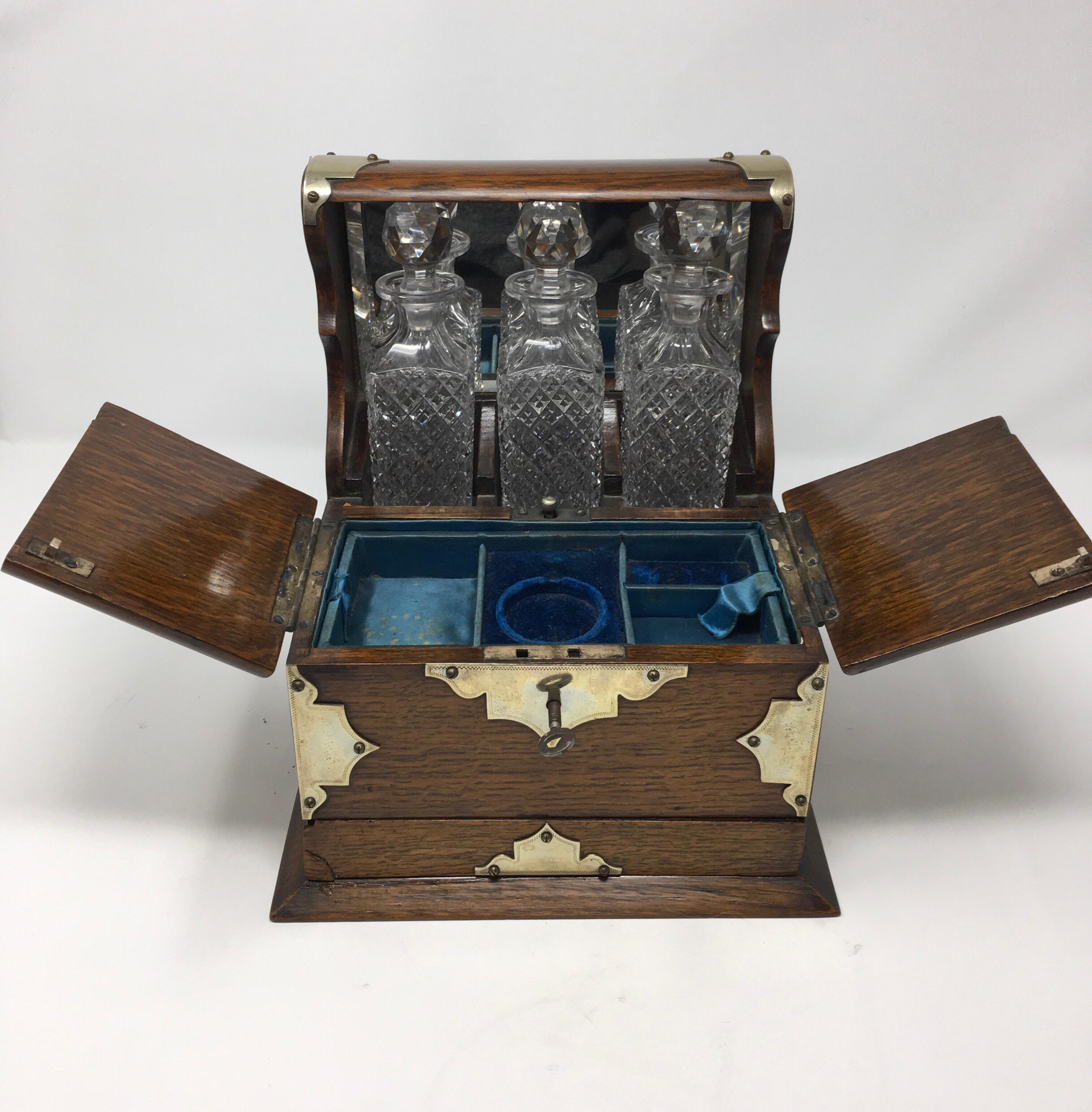 This is an antique English oak and brass Tantalus with three crystal decanters and a lower drawer. The three square crystal decanters are in good condition and each of them measure 1.75 inches wide, 1.75 inches deep and 6 inches high. The back of