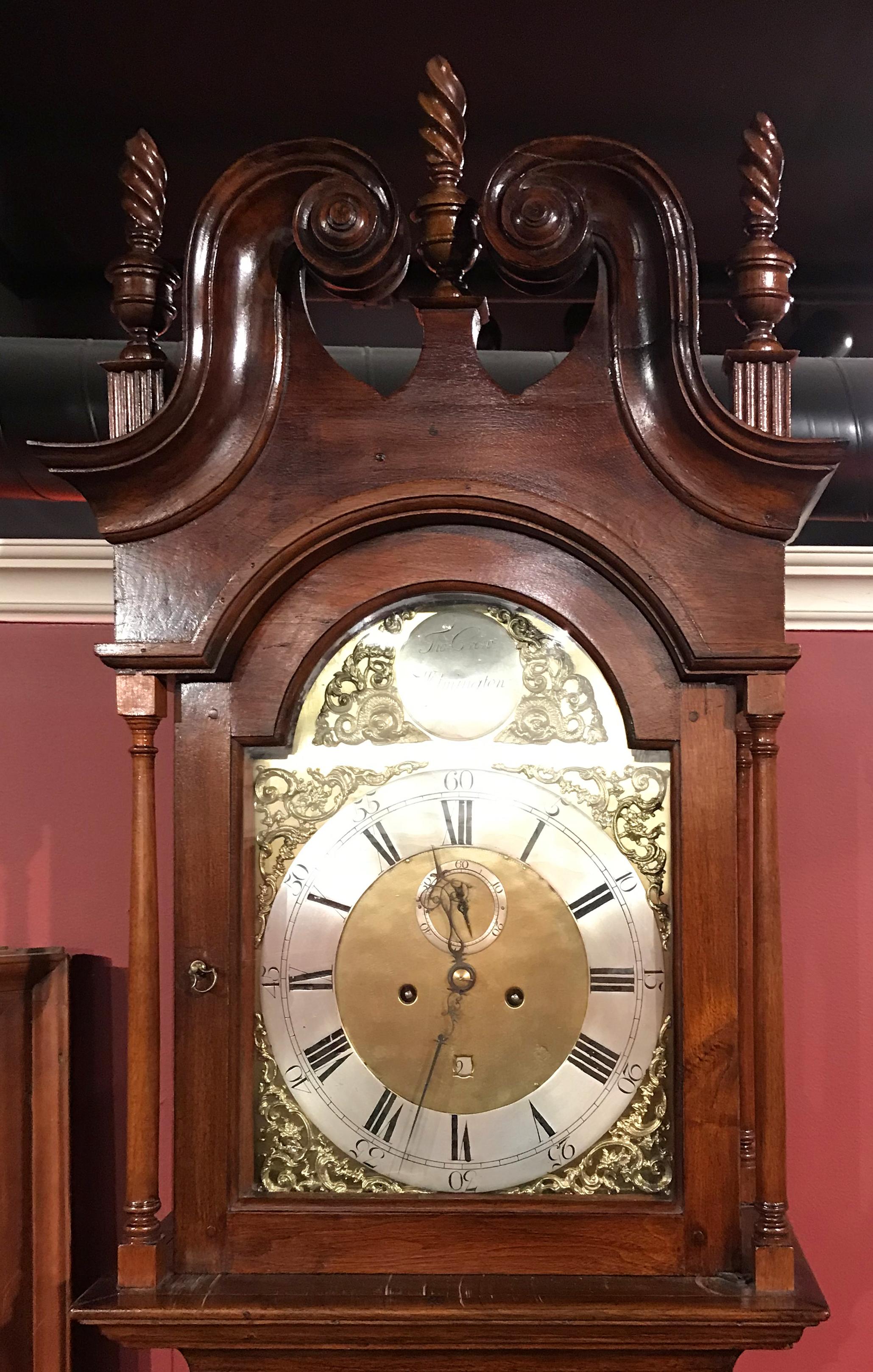 A fine mahogany tall case clock with carved swan neck broken pediment featuring three spiral form finials, its bonnet with a tombstone shaped glass door opening to a brass Roman numeral dial above which is inscribed “Tho. Crow, Wilmington,” pierced
