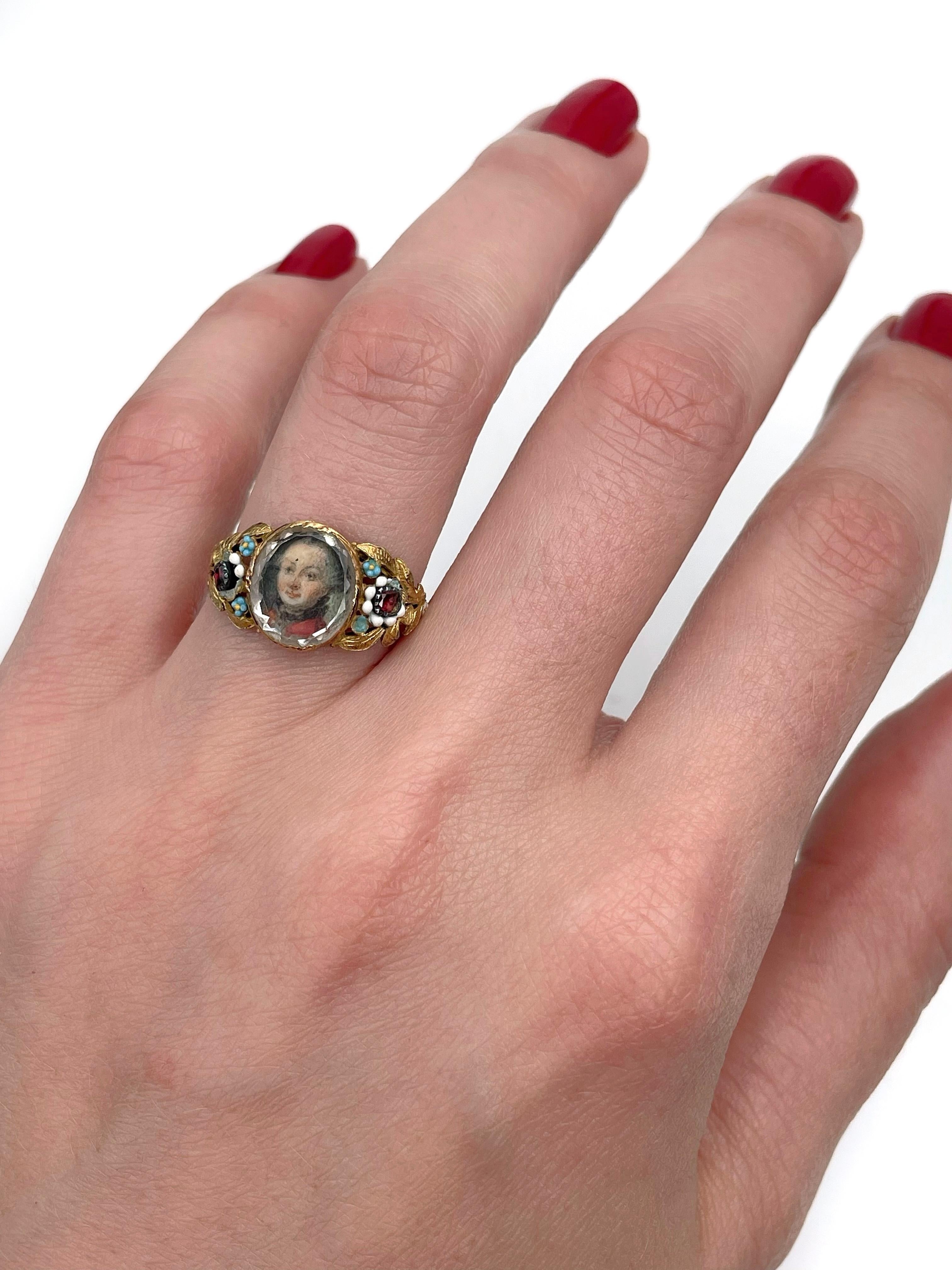This is a Georgian miniature portrait ring crafted in 18K yellow gold. The piece features a detailed hand painted portrait of a man (covered with the glass). 

The ring is rich in floral details. It is adorned with garnets and colourful enamel