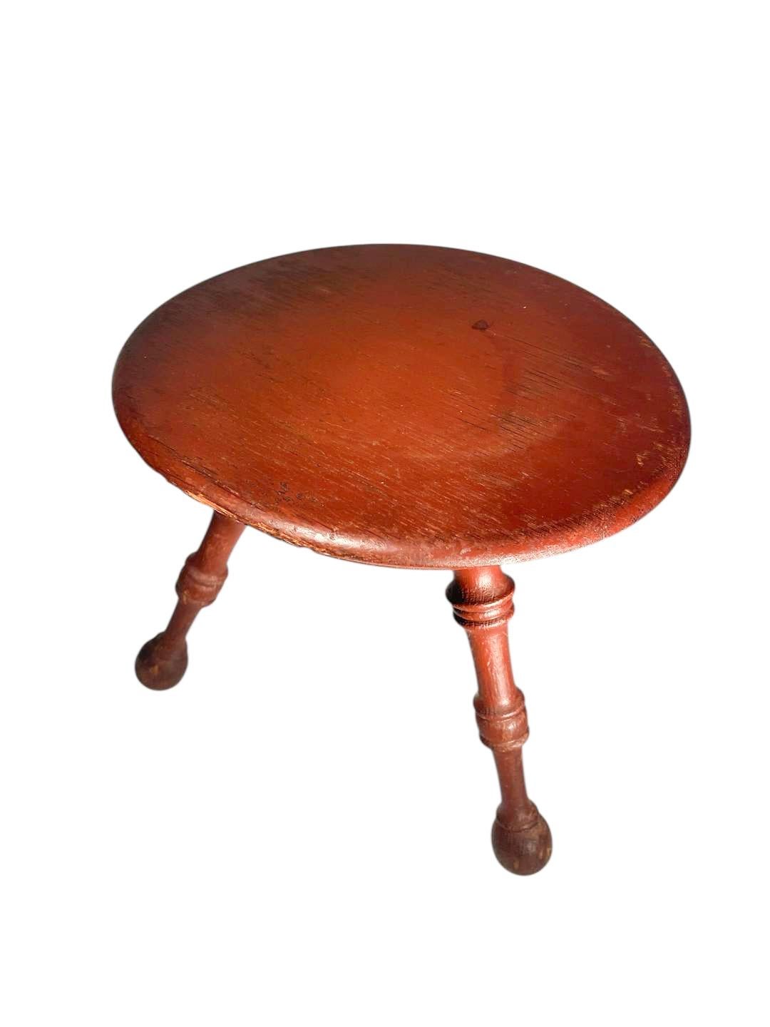 Boldly turned 18th century Windsor stool in old red paint having a slightly dished circular seat with molded sides raised on three ring and ball legs with ball feet.