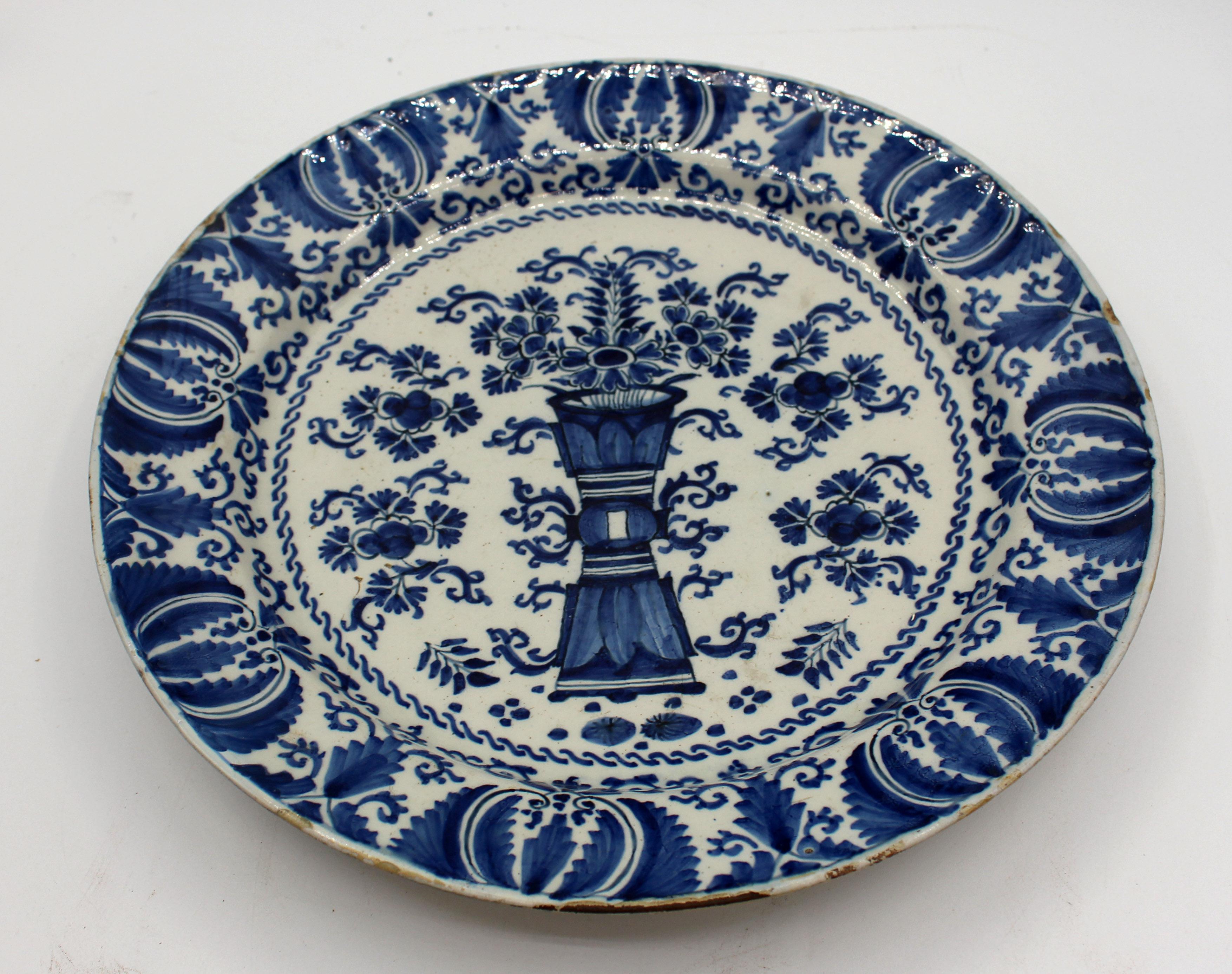 Late 18th century Delft blue & white chop plate. Central tall, pinched waist, cylindrical vase with flowers surrounded by flowers. Extensive alternating naturalistic motif border. Typical chips & frits for material, age & use. 11.75