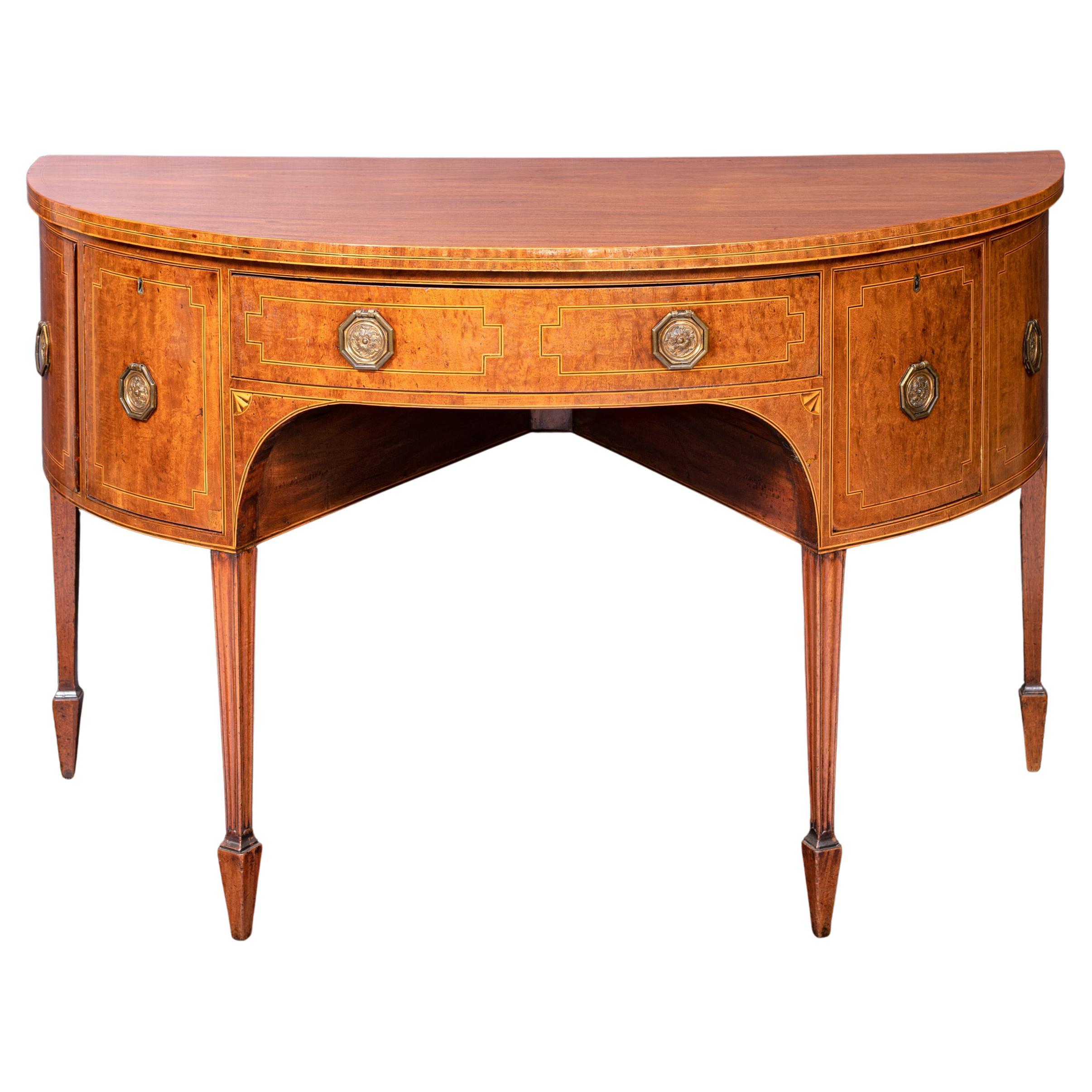 Ein feines georgianisches Mahagoni-Sideboard in Form einer Demi-Lune, mit einer zentralen Schublade, flankiert von zwei weiteren Schränken mit Messingringgriffen, auf spitzen Blockbeinen, die in kontrastierenden Spatenfüßen enden.

CIRCA