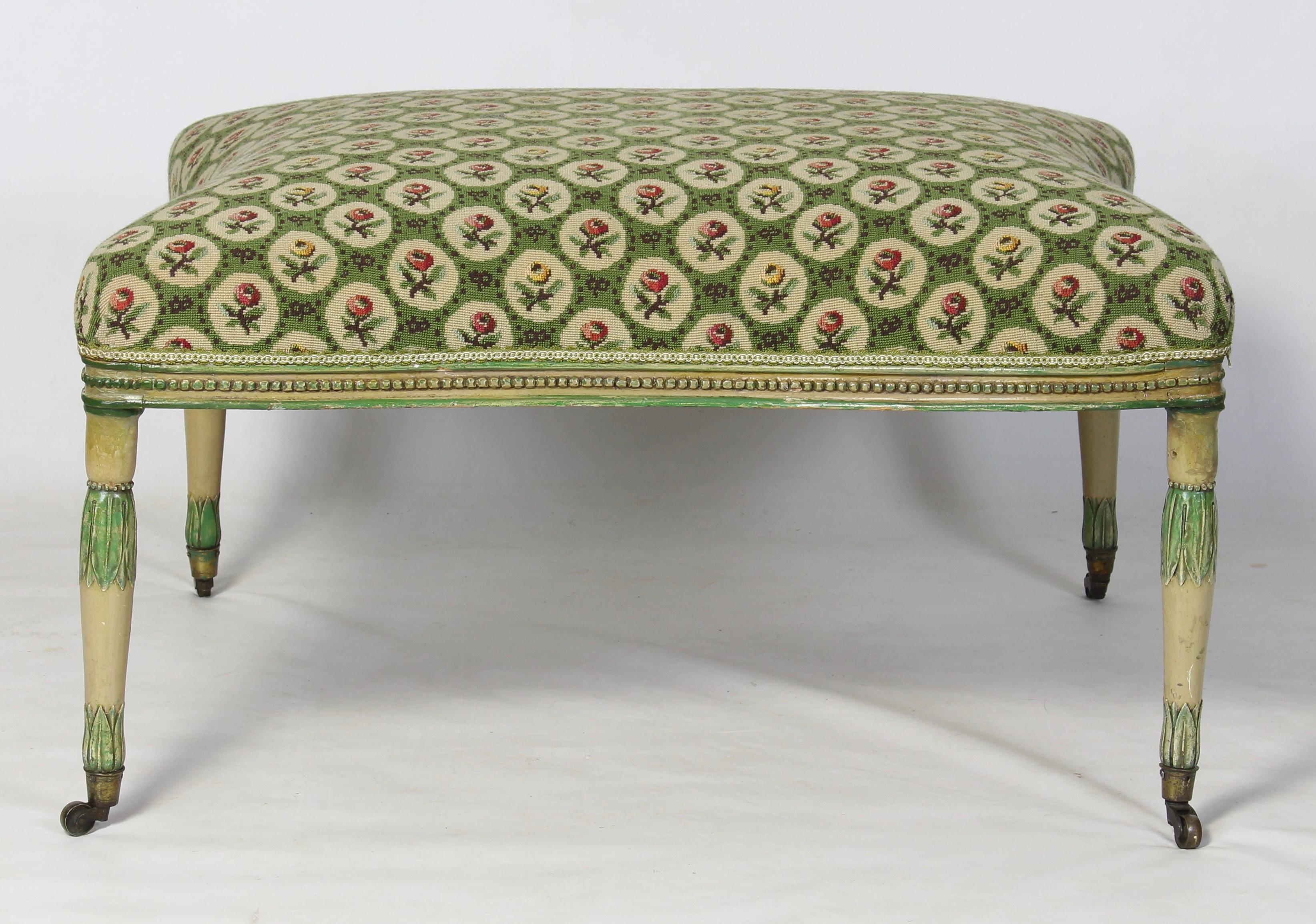 A large late 18th century English George III carved wood and paint decorated footstool or ottoman upholstered in green floral tapestry above four fluted tapering legs terminating in brass cup casters.