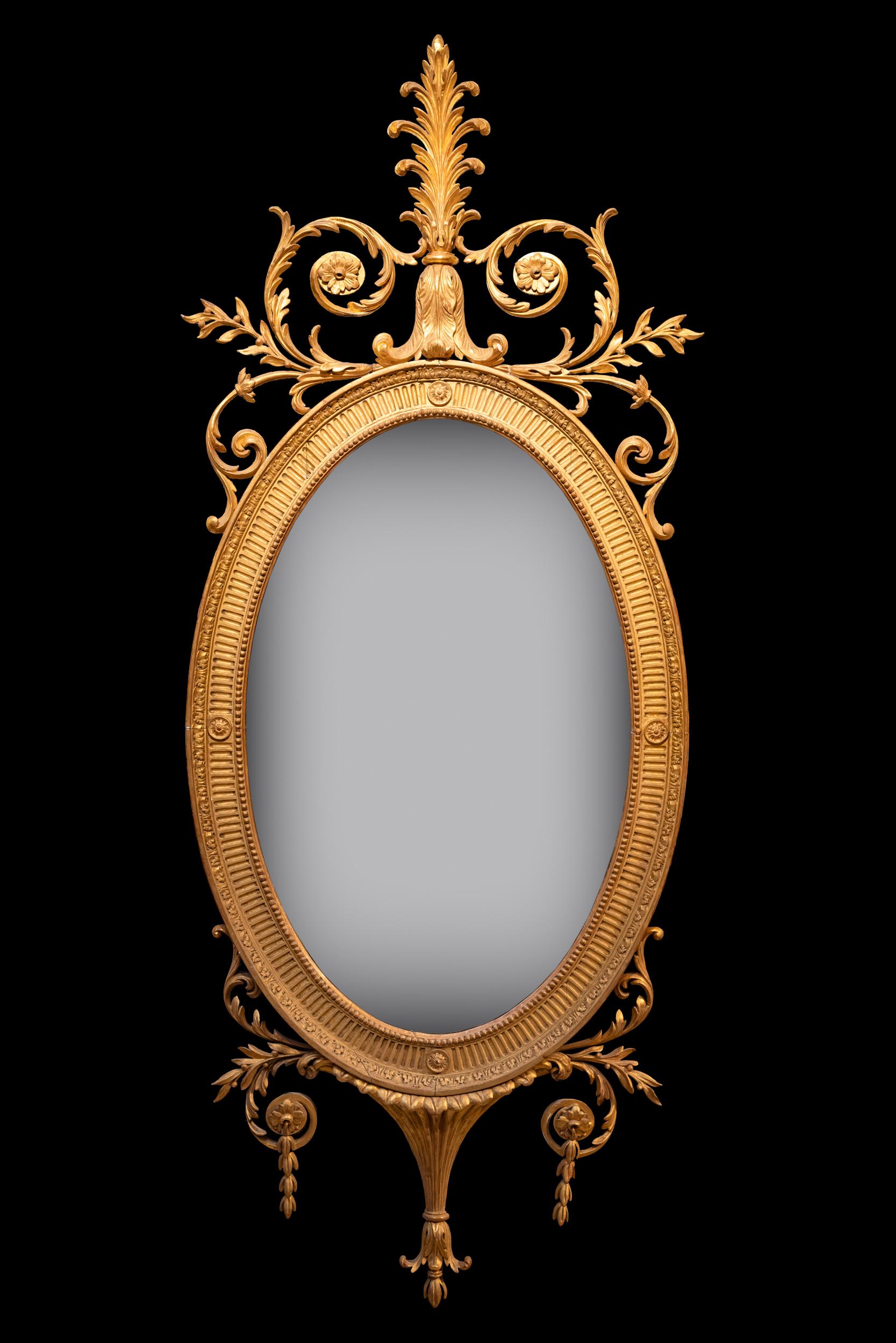 Très beau miroir mural / miroir de pilier ovale et doré du XVIIIe siècle, de style Adams. La plaque de verre de forme ovale est placée dans un cadre en bois doré mouluré et cannelé, sculpté à la main,  surmonté d'un écusson centré, minutieusement