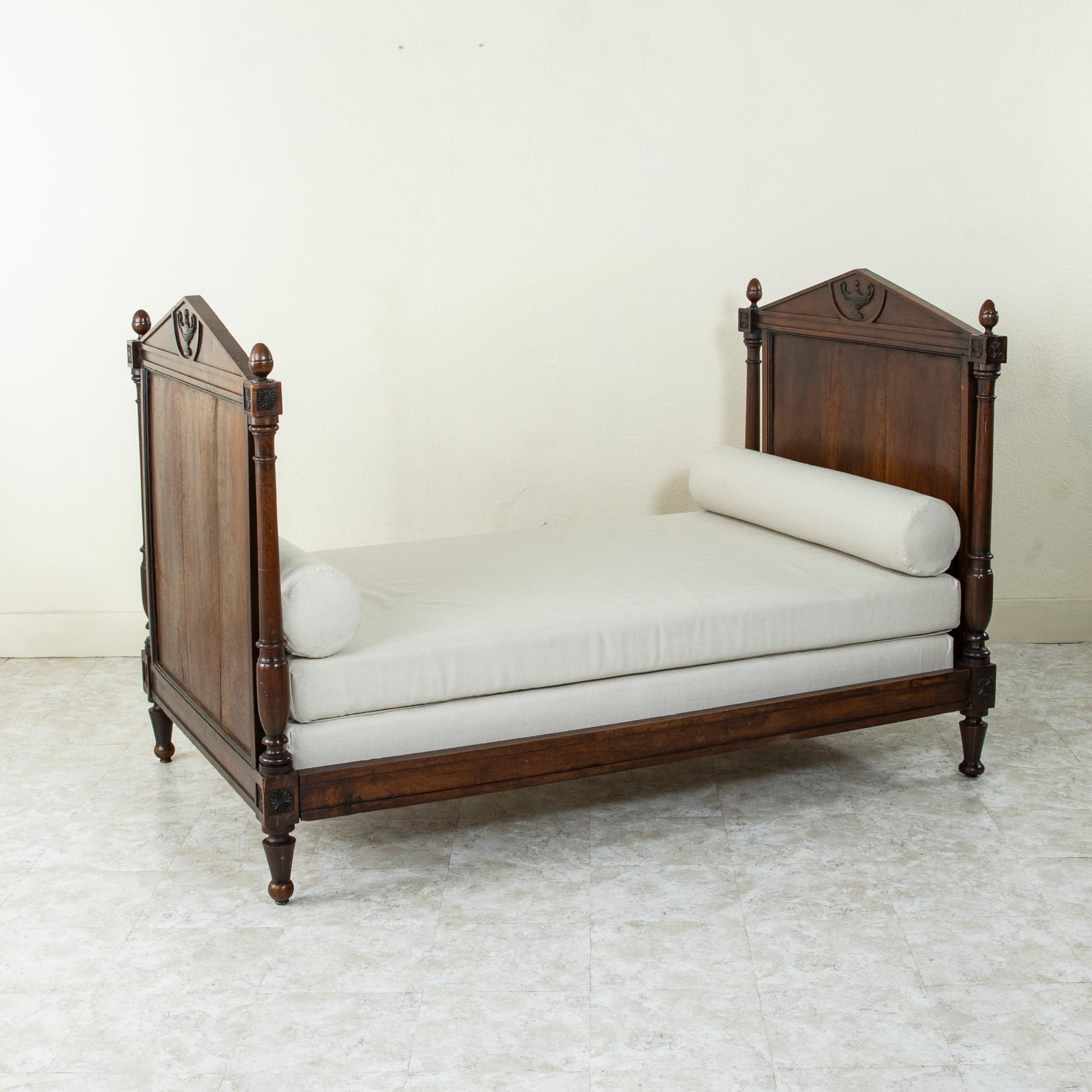 This French Directoire period walnut daybed from the late eighteenth century features hand carved rosettes on the die joints. Each end is crowned with a double faced pediment detailed with a hand carved central urn on each side. The mattress and
