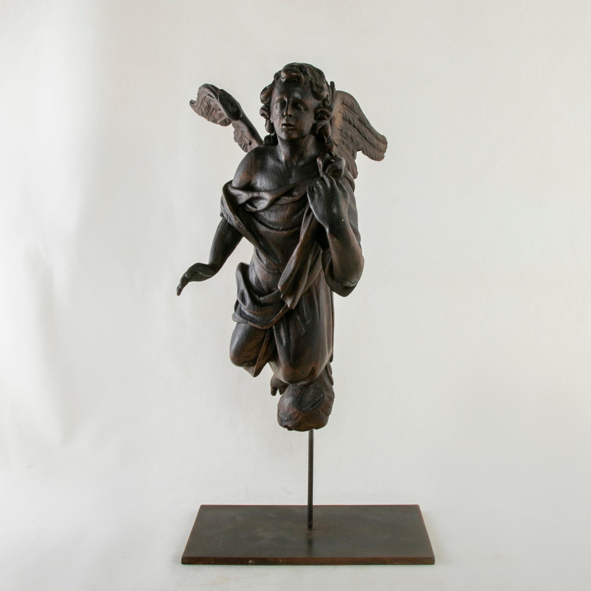 This hand carved angel sculpture from the late eighteenth century has wings outstretched as if in mid-flight. His left arm reaches up to his shoulder towards the cloth draped around him. Suspended from eyelets on his back, this sculpture is mounted