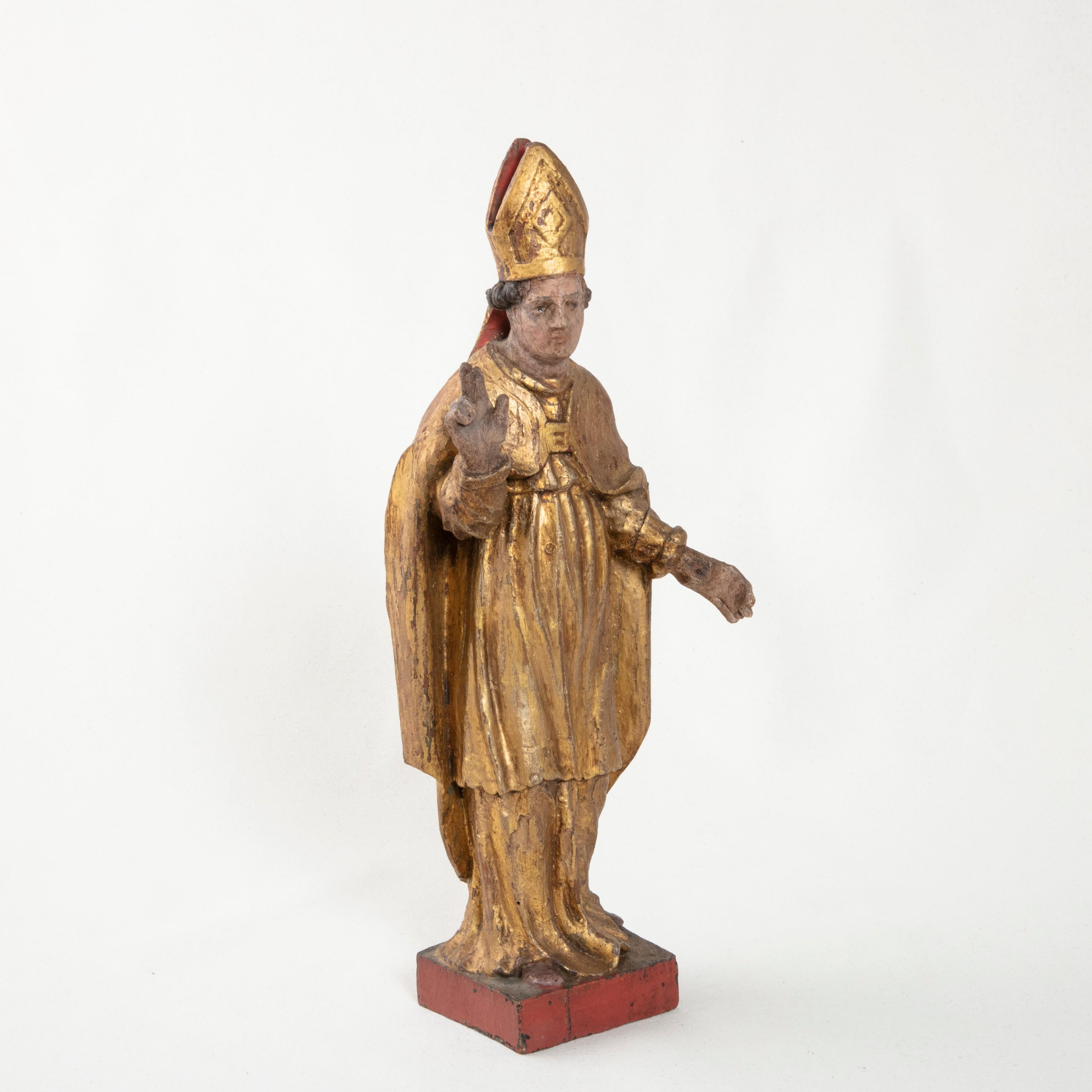 This late 18th century French hand carved giltwood polychrome statue is of a bishop dressed in golden robes. On top of his head rests a typical bishop's gold mitre hat with a red underside. His right hand is held outstretched to offer benediction.