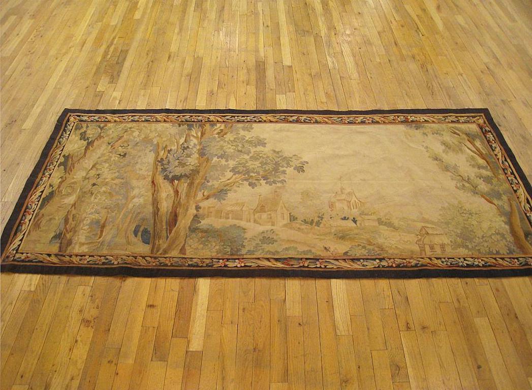 A French landscape tapestry from the late 18th century, envisioning a series of cottages on verdant hills at center, flanked by large stately trees to the left and right. Enclosed within a scrolling border of flowerheads and acanthus leaves. Wool