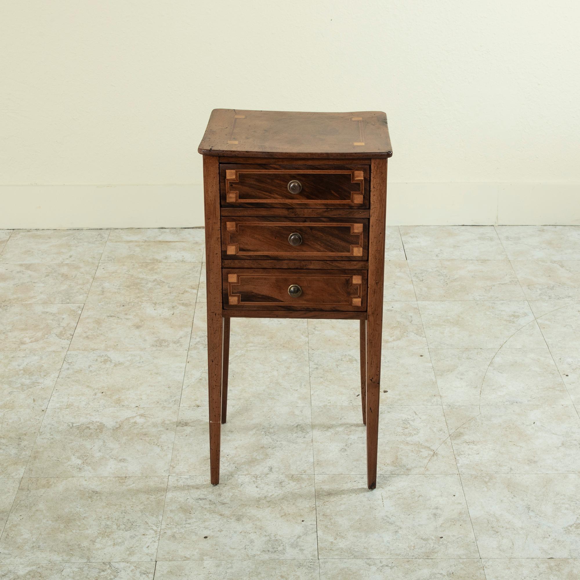 From the city of Lyon, this late eighteenth century French Louis XVI period cherrywood nightstand or side table features inlaid lemonwood and walnut that create an inset border with indented corners on the top and drawer fronts. Its three drawers of