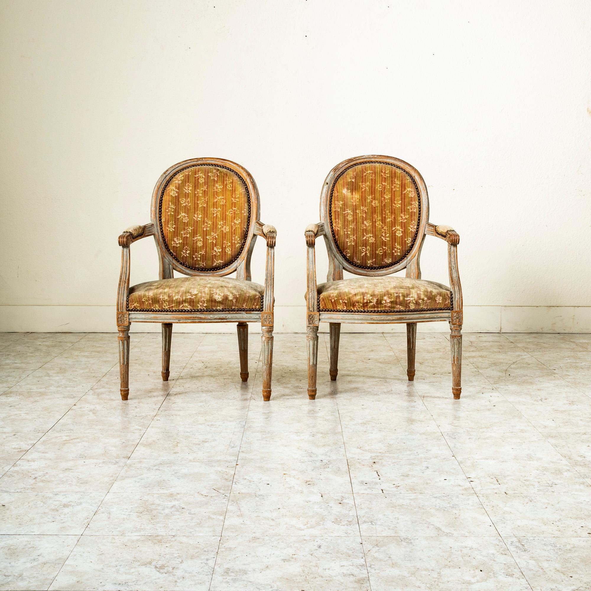 This pair of late eighteenth century French Louis XVI period armchairs features its original paint worn to a beautiful patina that only comes with age. This classic pair features medallion backs, fluted arms with scrolled armrests, and tapered legs