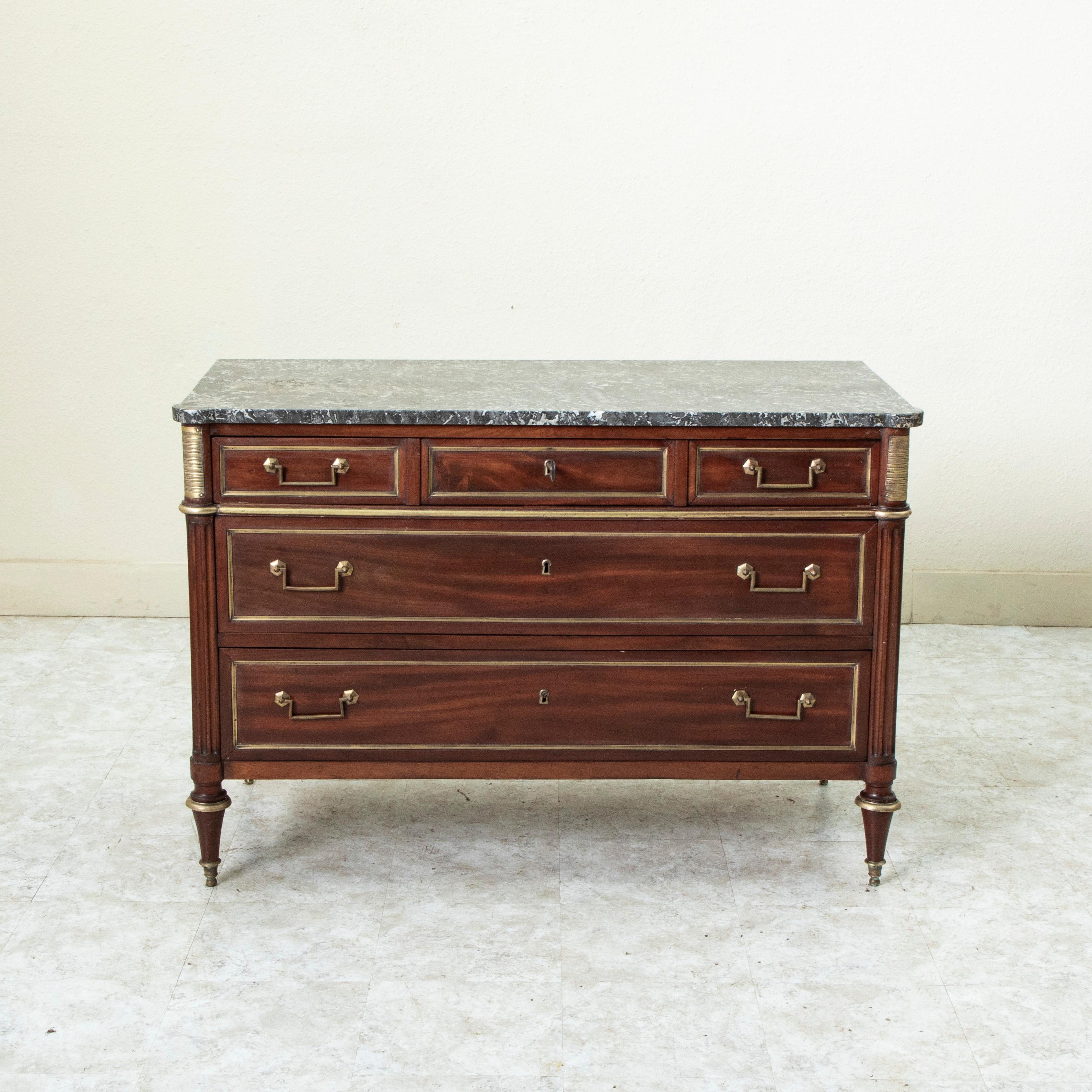 This late eighteenth century French Louis XVI period mahogany commode or chest of drawers features a beveled Saint Anne marble top. The corners are detailed with fluting and striated bronze plaques. Each panel is surrounded by a bronze border, and