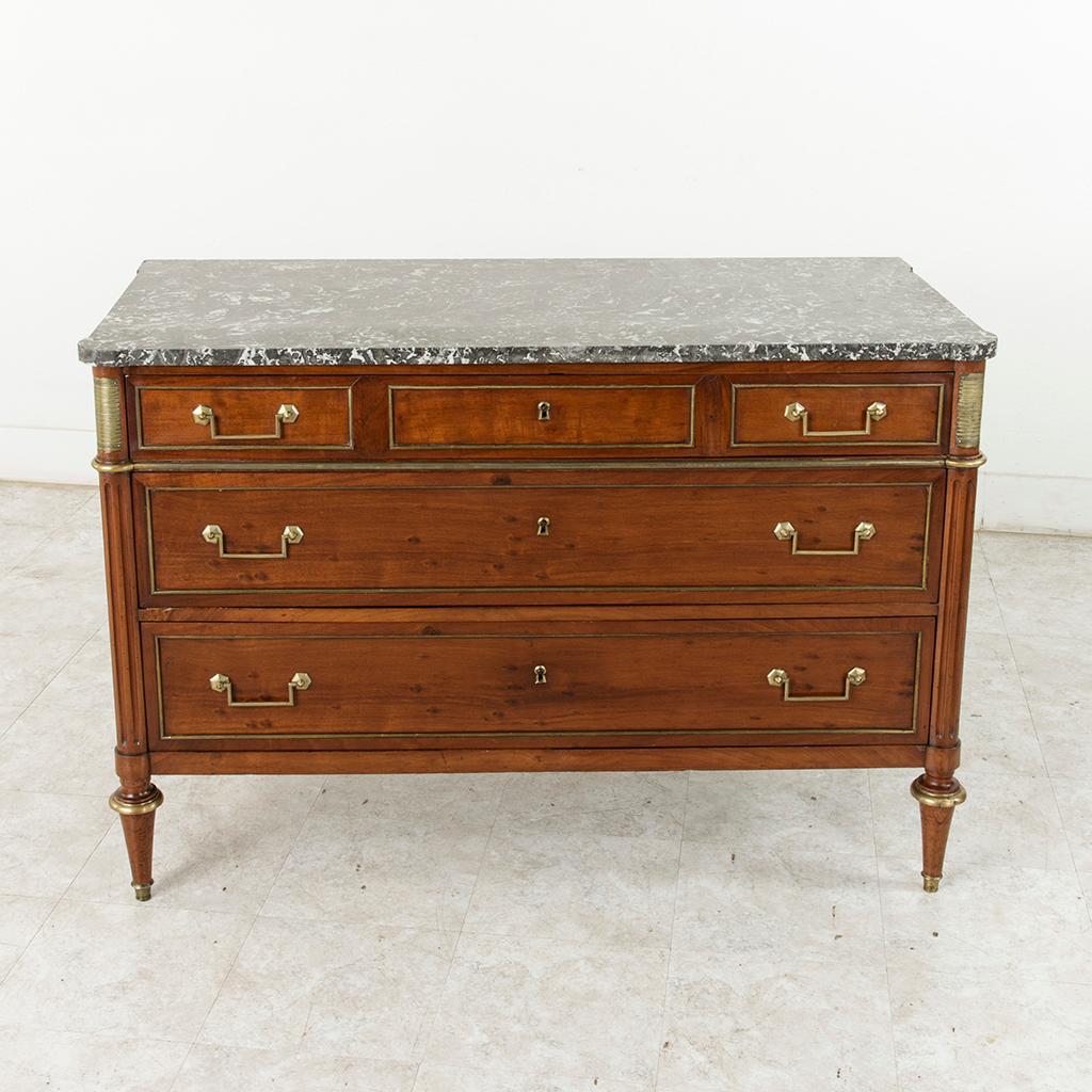 This late 18th century French Louis XVI period mahogany commode or chest features a Saint Anne marble-top and striated bronze plaques on the rounded front corners. Bronze banding lines the panels and drawer fronts of the chest. Its three drawers of