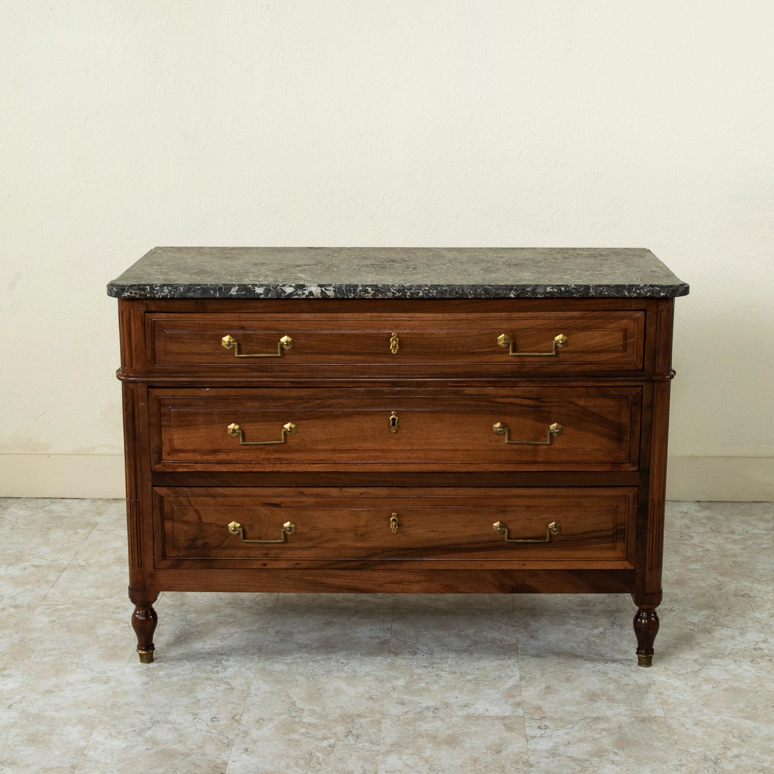 This late 18th century French Louis XVI period walnut commode or chest of drawers features a beveled Saint Anne marble top and fluted rounded corners. Its three drawers of dovetail construction are fitted with classic squared drop handle drawer