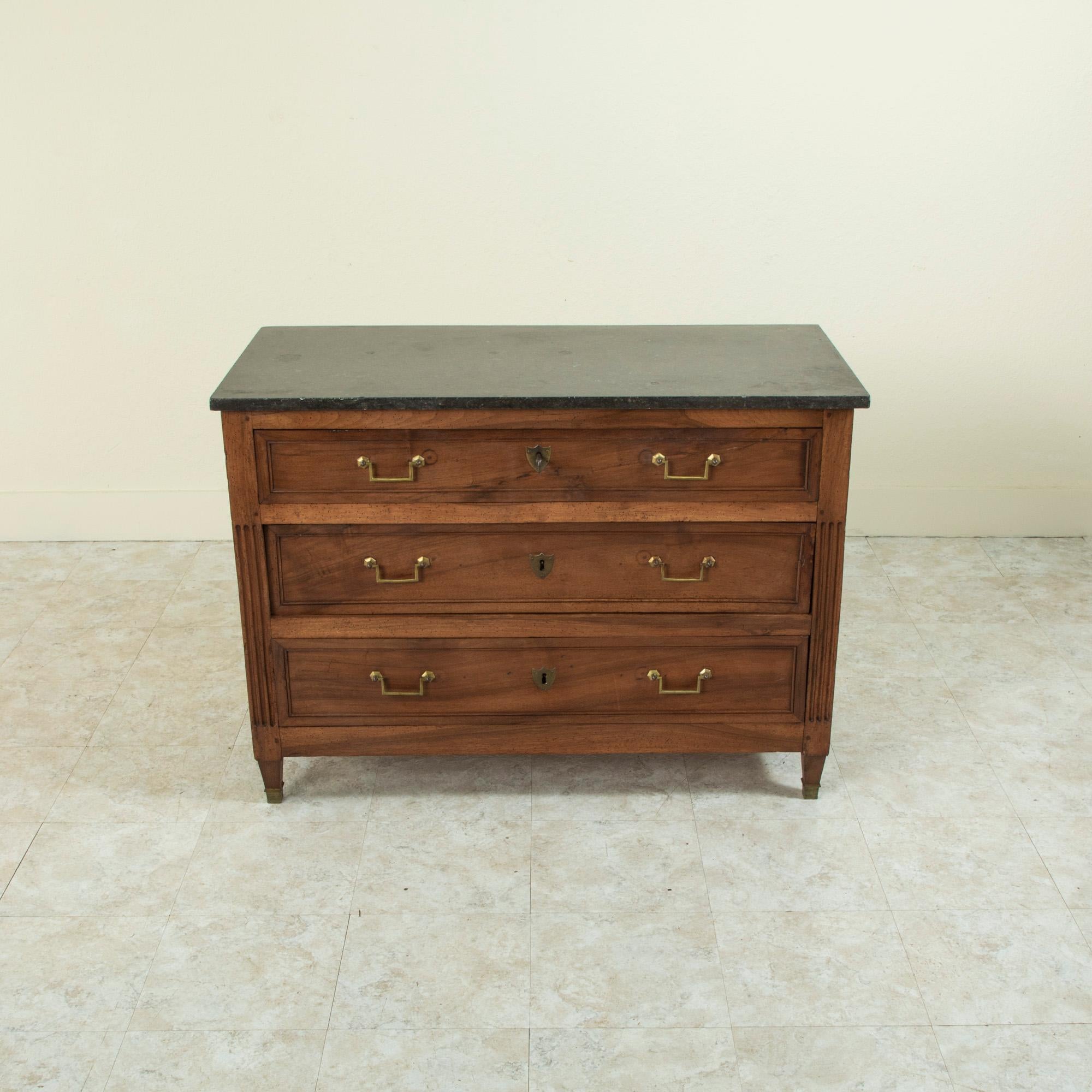 This late eighteenth century French Louis XVI period walnut commode or chest of drawers features a black marble top. The corners are detailed with hand carved fluting and terminate in tapered square legs finished with bronze sabots. Its three