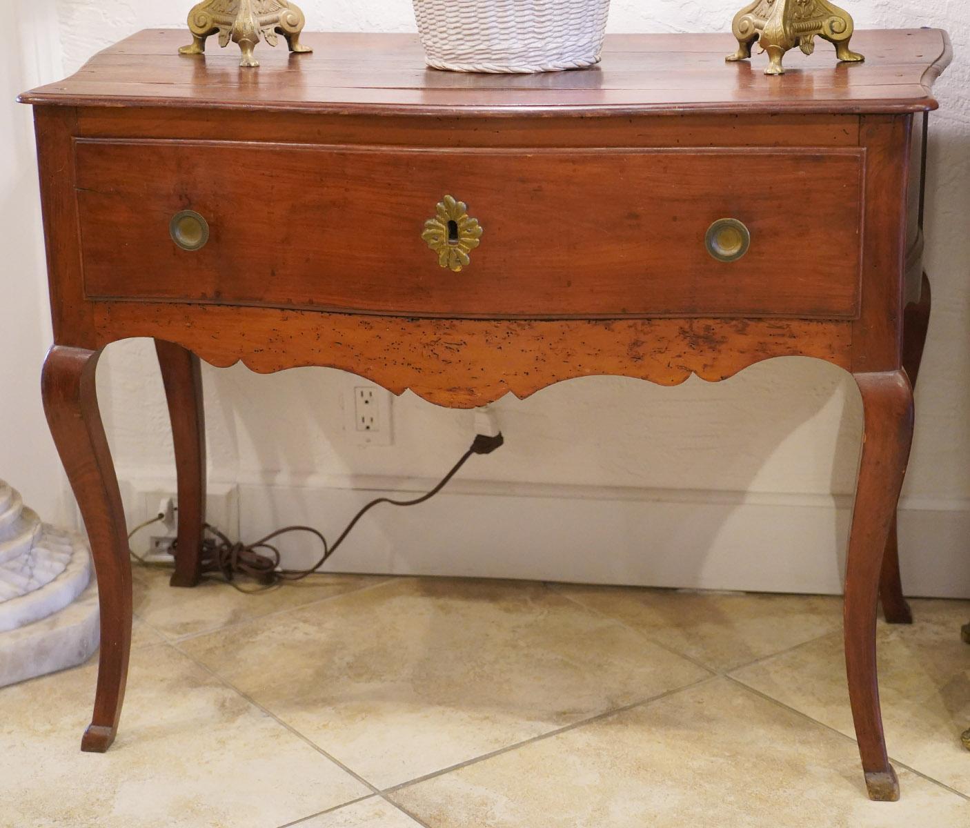 This late 18th century French Provincial cherry serpentine console table features a shaped top above one long serpentine front drawer retaining original hardware and a carved scalloped apron. The table rests on four cabriole legs ending in carved