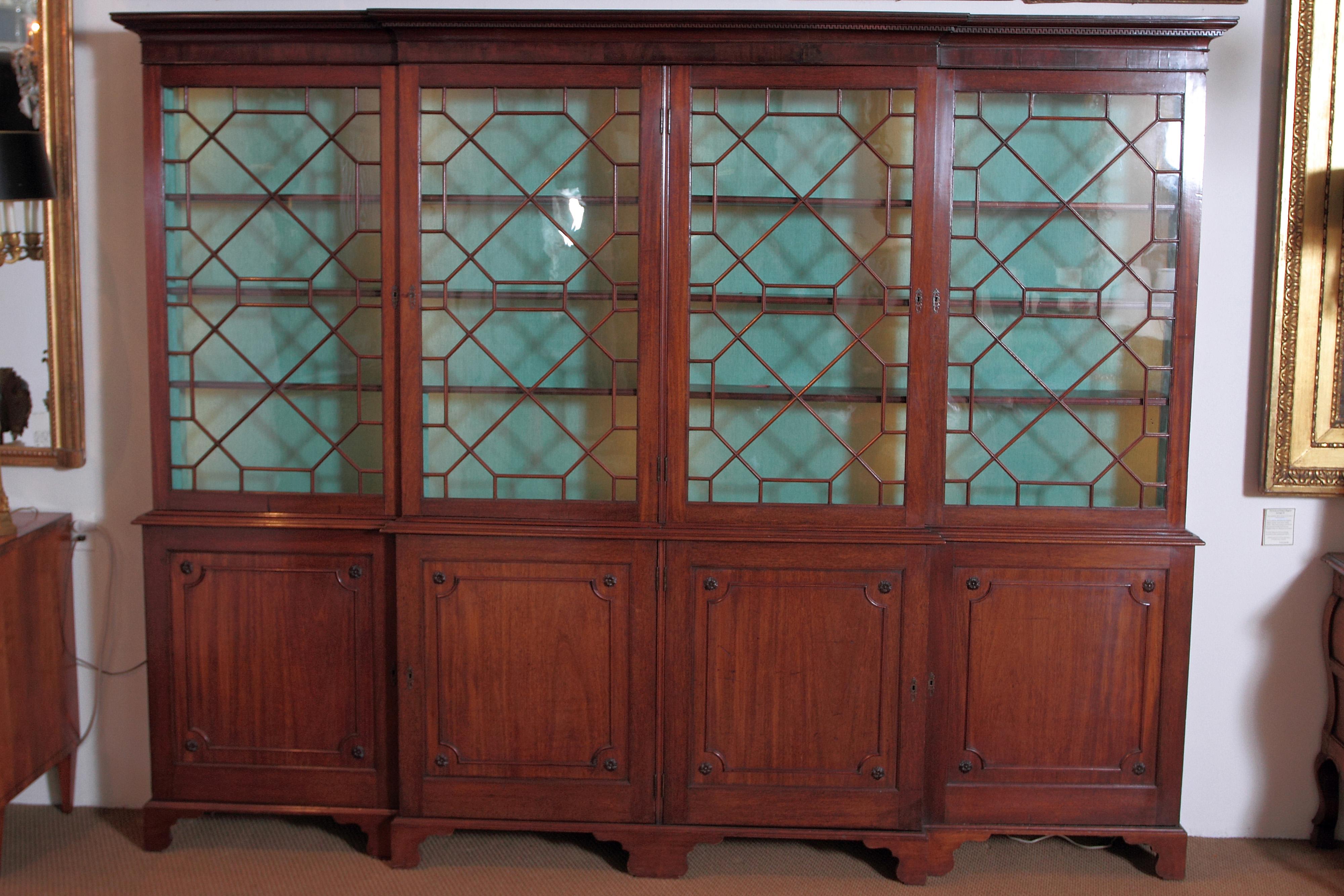 A large Georgian mahogany breakfront bookcase with carved Greek Key trimmed cornice above four mullioned glass doors. The interior is lined with green fabric and has dimmable lighting. Each section has three fixed wooden shelves. Below are four