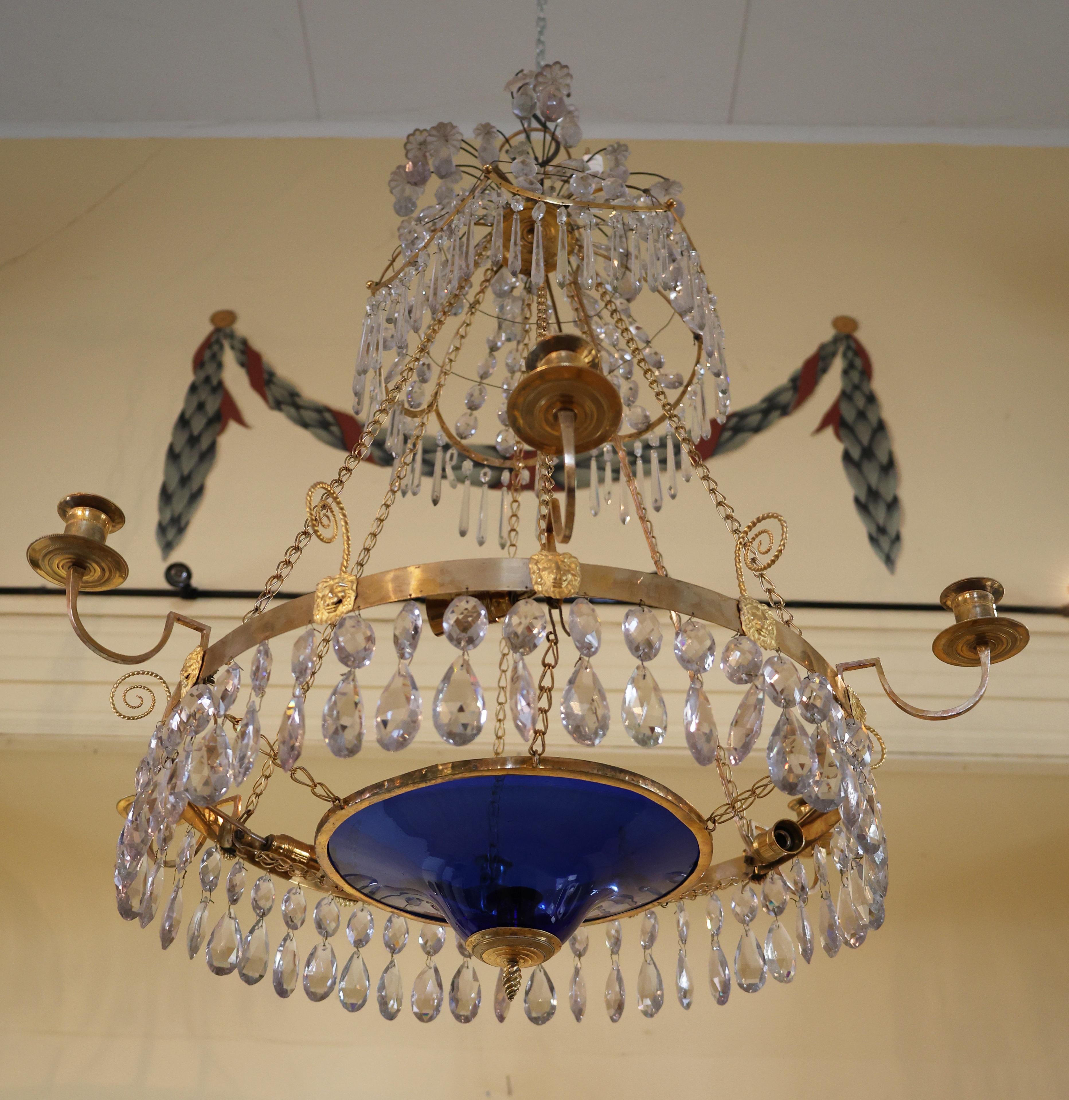 Lustre de la fin de la période gustavienne avec une coupe bleue, fabriqué à Stockholm dans les années 1790. L'anneau central doré et bruni est décoré de sculptures de masques de lion. L'anneau comporte six bras légers et six chaînes le relient à la