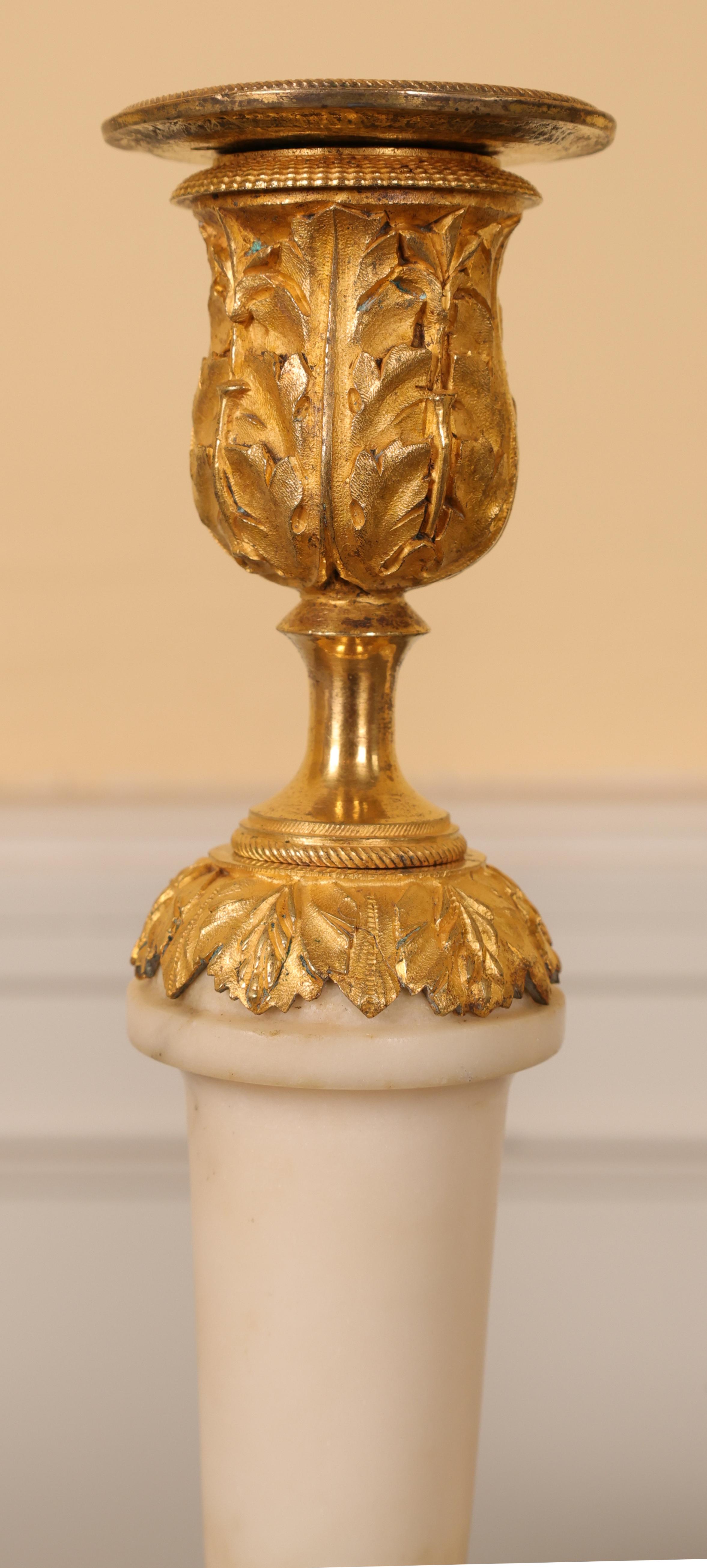 Coppia di candelieri francesi Luigi XVI della fine del XVIII secolo. I fusti e le basi sono in marmo bianco di Carrara, mentre gli accessori sono in bronzo dorato brunito. La base è decorata con drappeggi e nappe, mentre le prese di luce presentano
