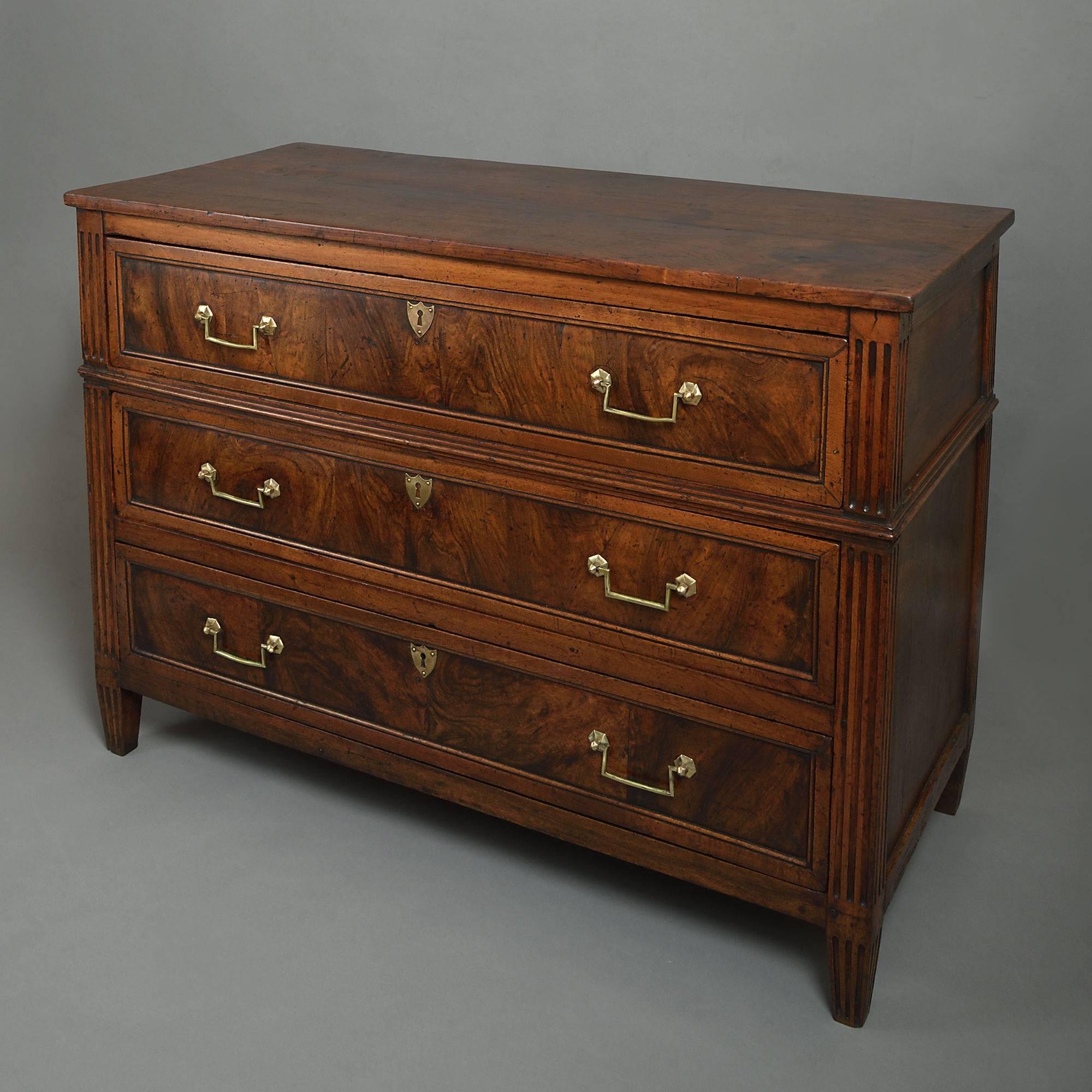 A good late 18th century Louis XVI Period walnut commode, the shaped cleated wooden top above three panelled drawers with rectangular brass handles, the corners with fluted carving, all set upon turned tapering legs.