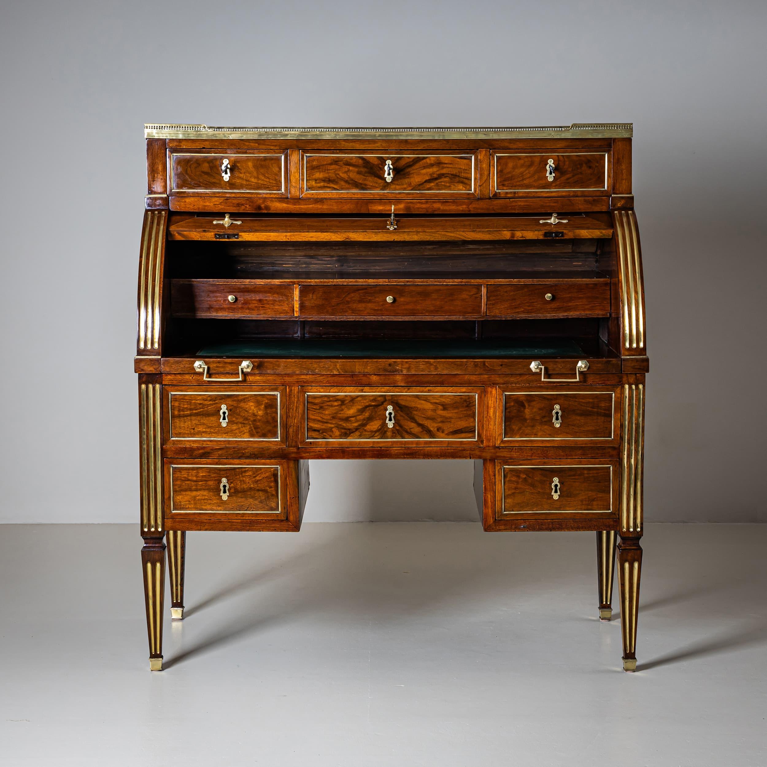French roll-top desk resting on fluted legs with brass inlays and sabots. The refined fluting continues along the pilasters, creating a cohesive design. The lower section offers ample storage with five drawers, each framed by delicate brass