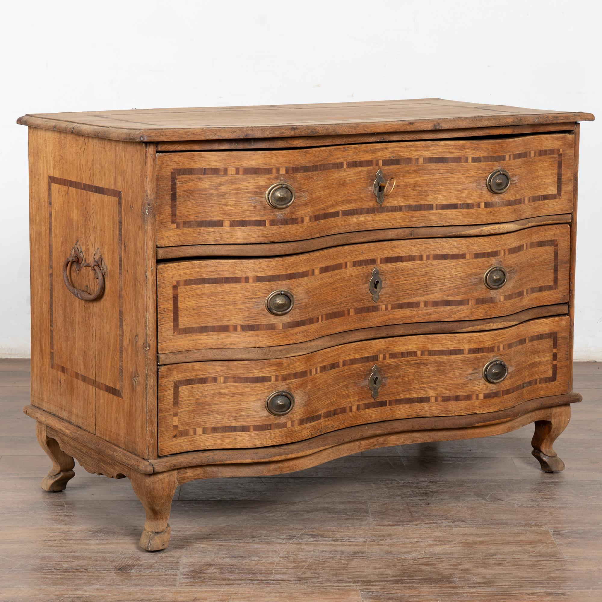 This antique rococo large oak chest of drawers is accented with contrasting inlaid trim along the top, sides and three drawer fronts. It features a serpentine front, iron side handles, brass drawer pulls and is raised on cabriolet feet. 
This