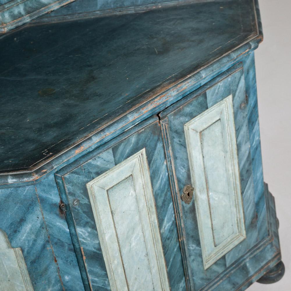 An 18th century two part Swedish painted corner cupboard finished in blue veined marble effect in contrasting blues. The cabinet is set upon bun feet and has its original furnishings and shelving to the interiors.