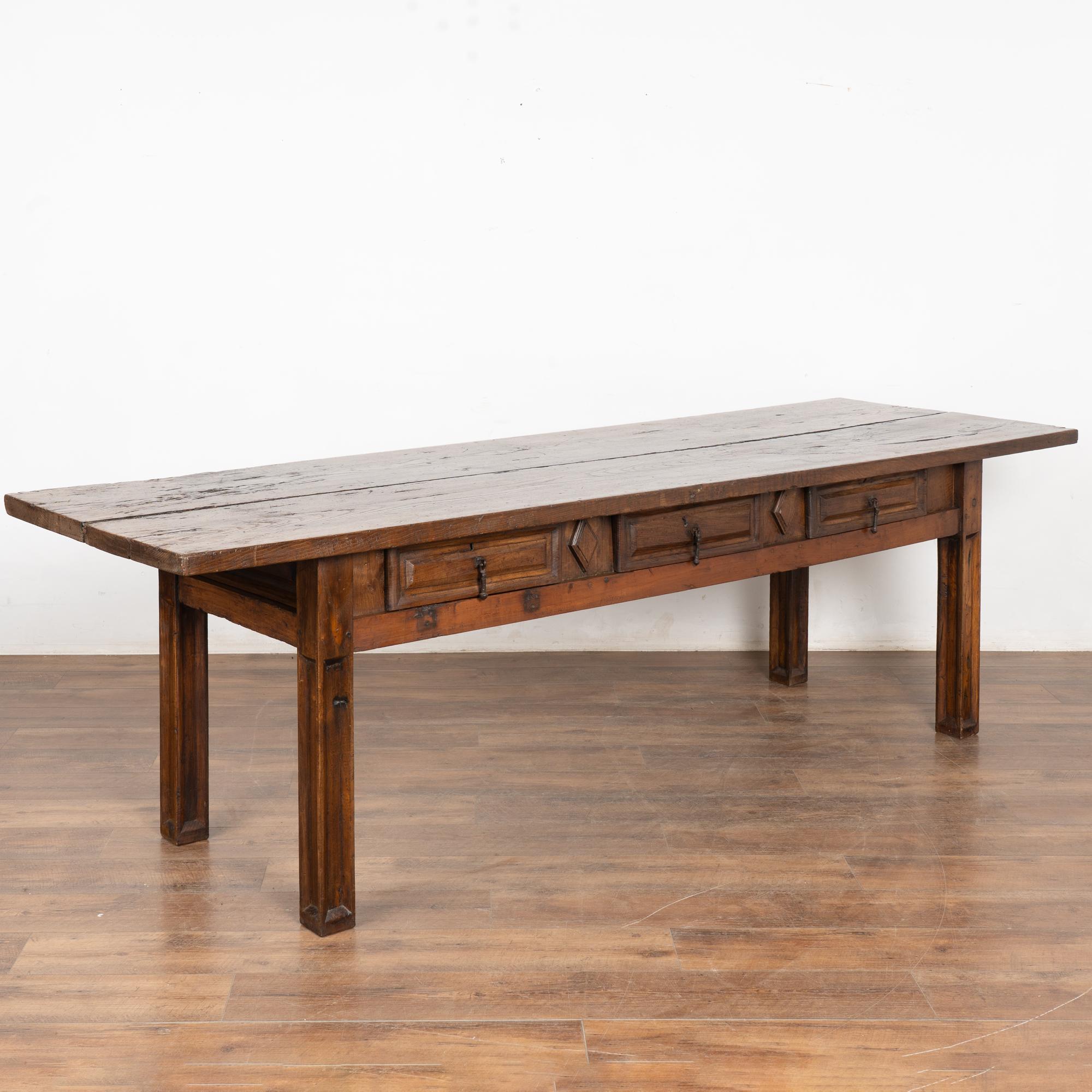 Dark oak console table with three drawers resting on four squared legs. Simple carved panels along drawer fronts, each with metal pull. Carved diamond accents between the drawers. 
Please enlarge photos (especially of top) to appreciate the dark