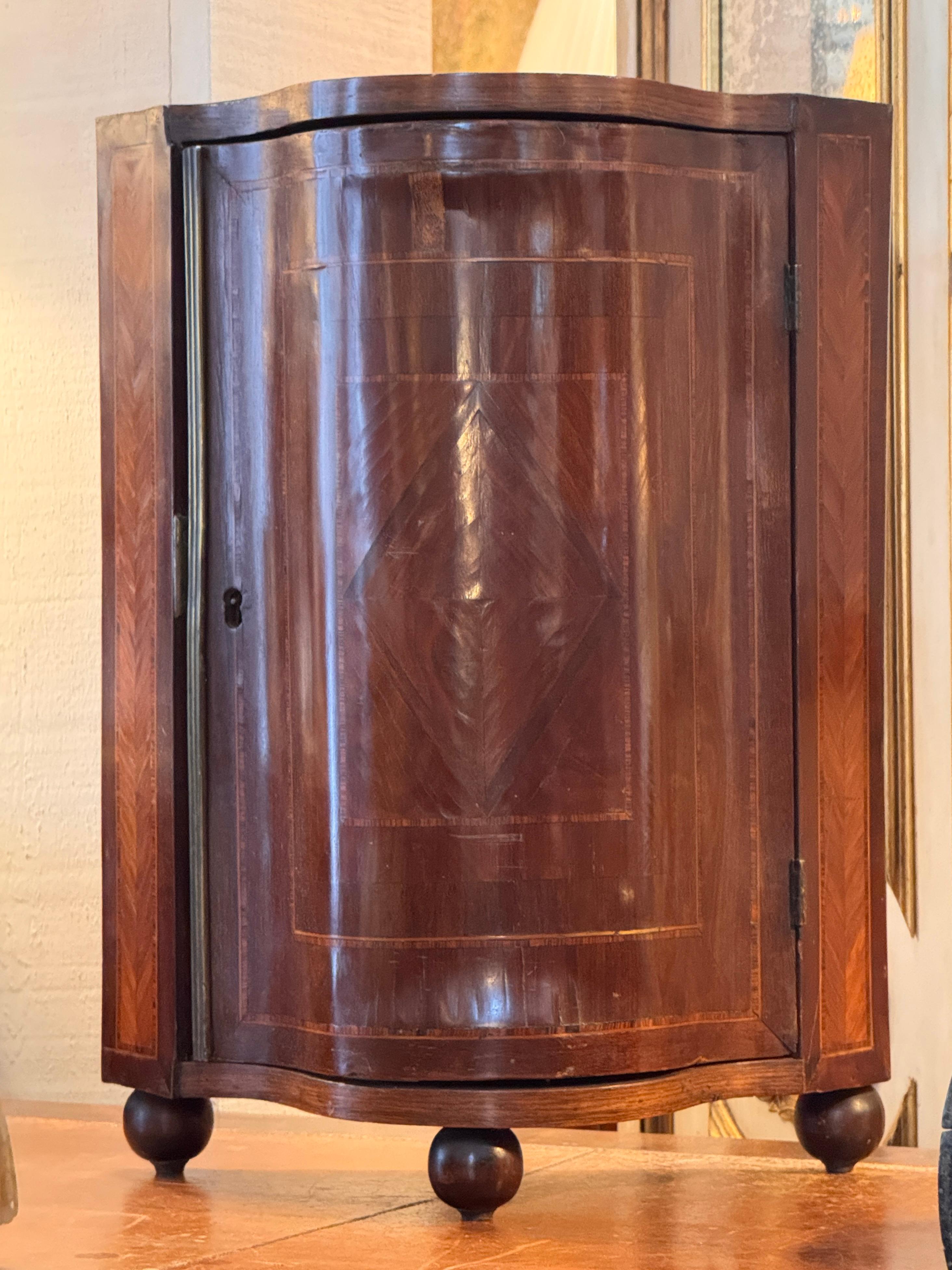 Italian miniature cabinet with a shaped front and finely inlaid veneers, including chevron-pattern reserves on the sides and banded borders framing the door. The curved interior shelf follows the outline of the case, and the piece stands on turned
