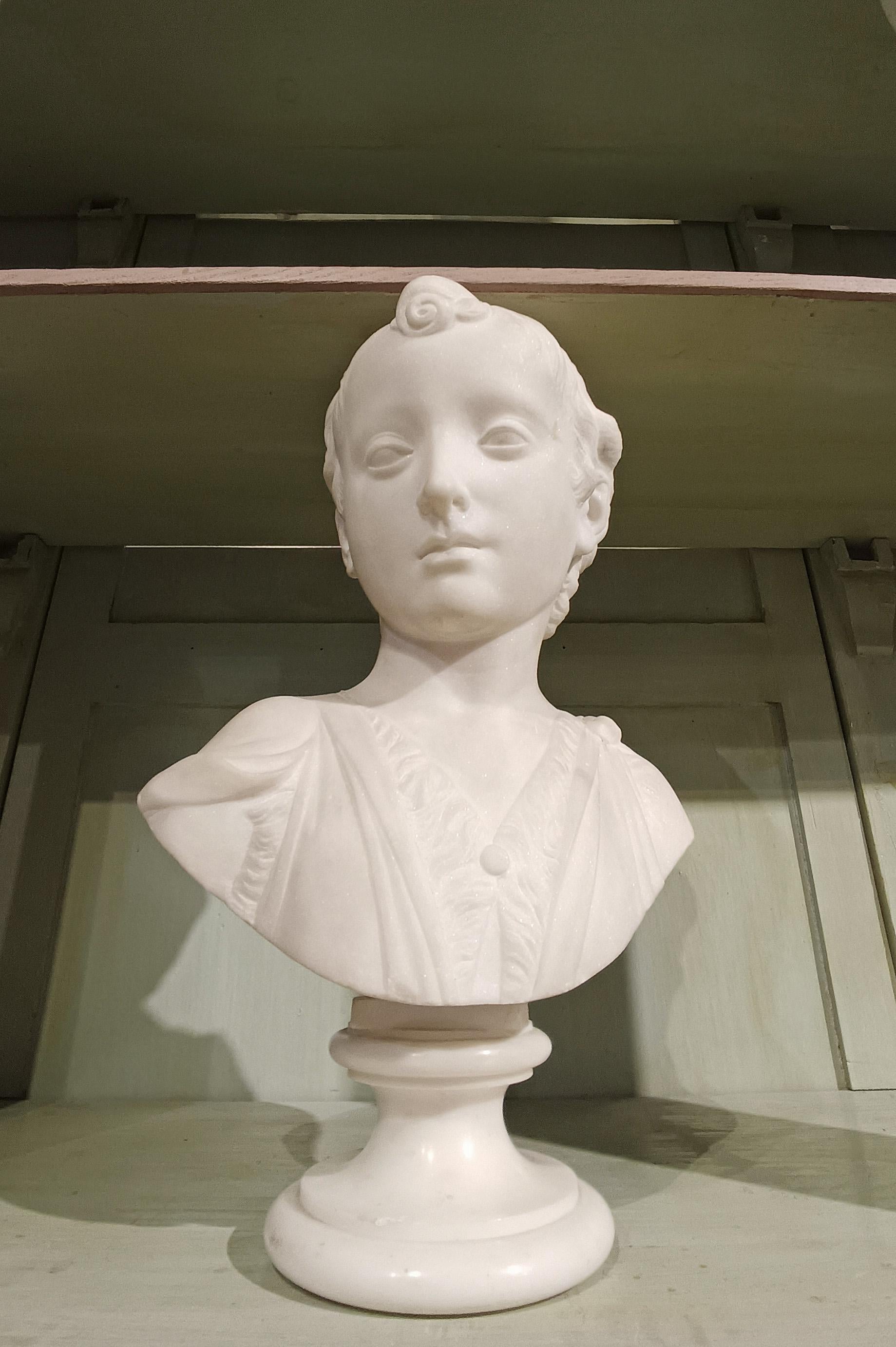 Elegante busto de mármol blanco, sostenido por una base circular de mármol torneado. El busto representa a un joven auriga, retratado con expresión serena y el rostro enmarcado por una cabellera rizada y voluminosa. El chitón, la túnica tradicional