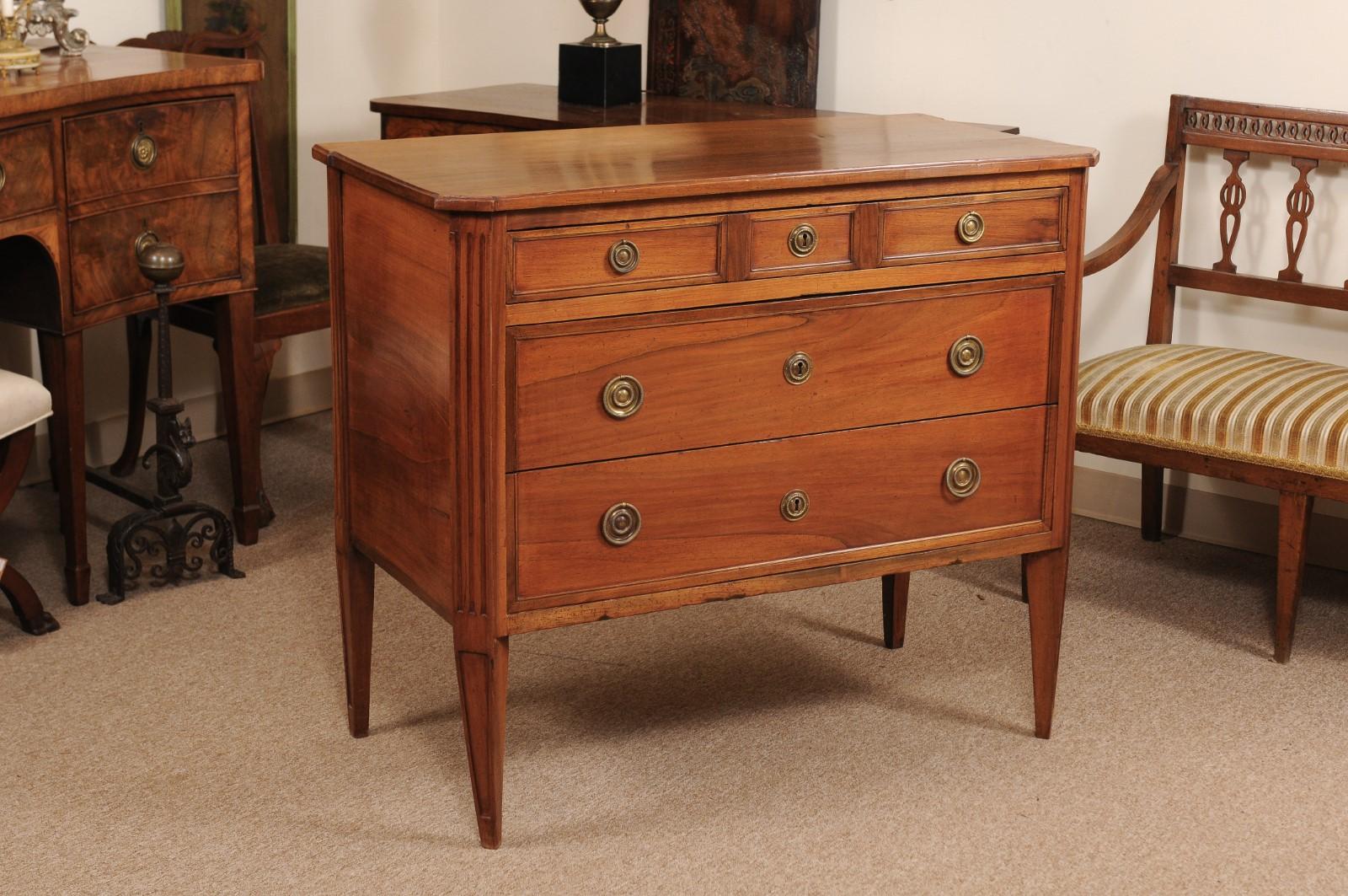 The late 18th century Louis XVI commode in light walnut with canted corners, fluted carving and 5 drawers terminating in tapered legs.