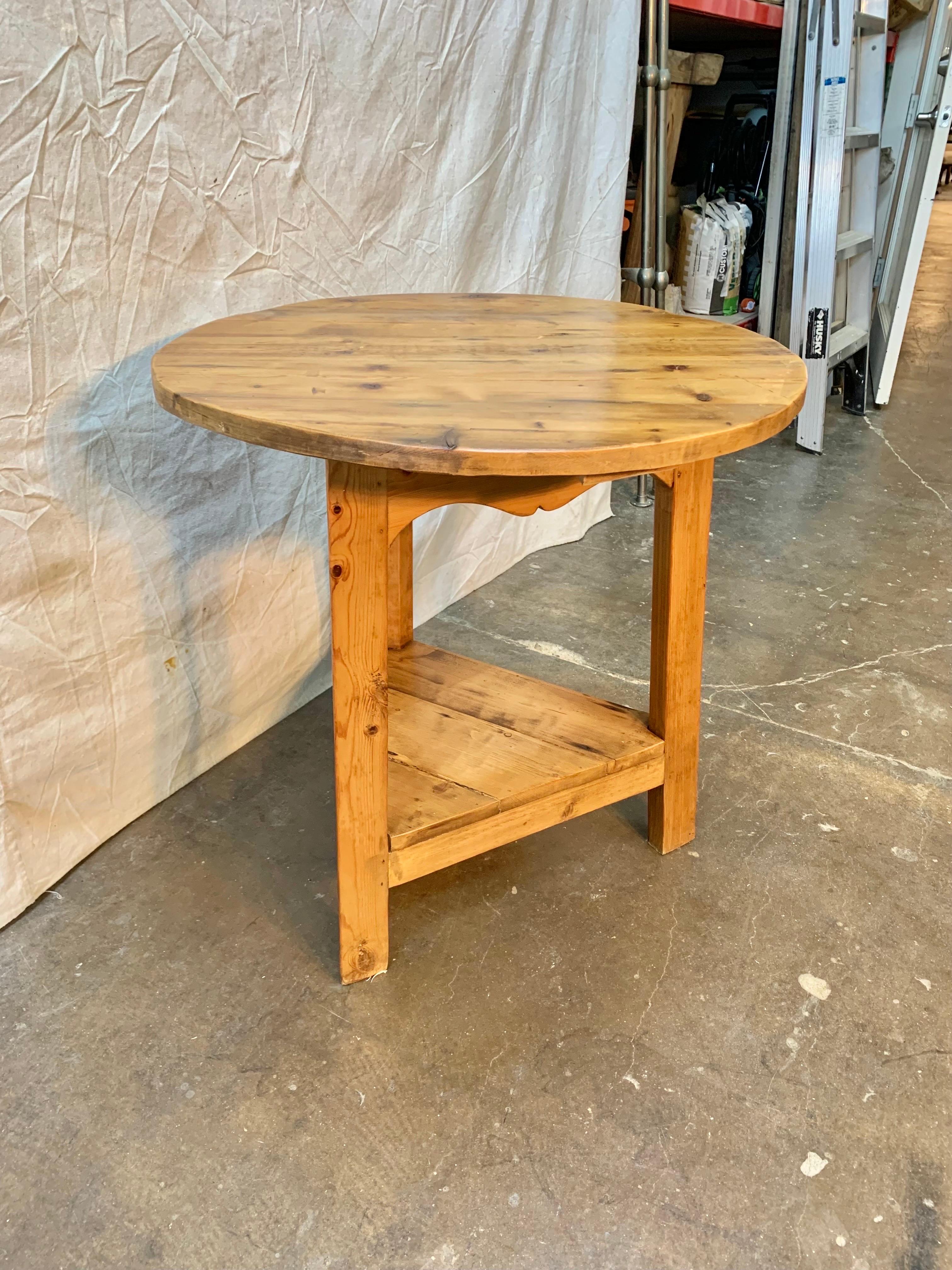 This English Cricket Table was crafted in the late 1900's from old growth pine. The piece features a circular top resting on three angled legs that have been adjoined with a triangular lower shelf. Cricket tables are in essence English pieces of
