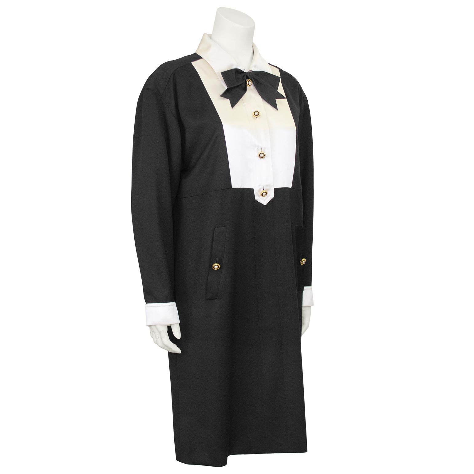 Vestido de esmoquin Chanel by Karl Lagerfeld de finales de los años 80. El vestido tiene cuello, pechera y puños de seda blanca marfil con un lazo de seda negra y botones metálicos dorados. El resto del vestido es de lana negra en contraste, con