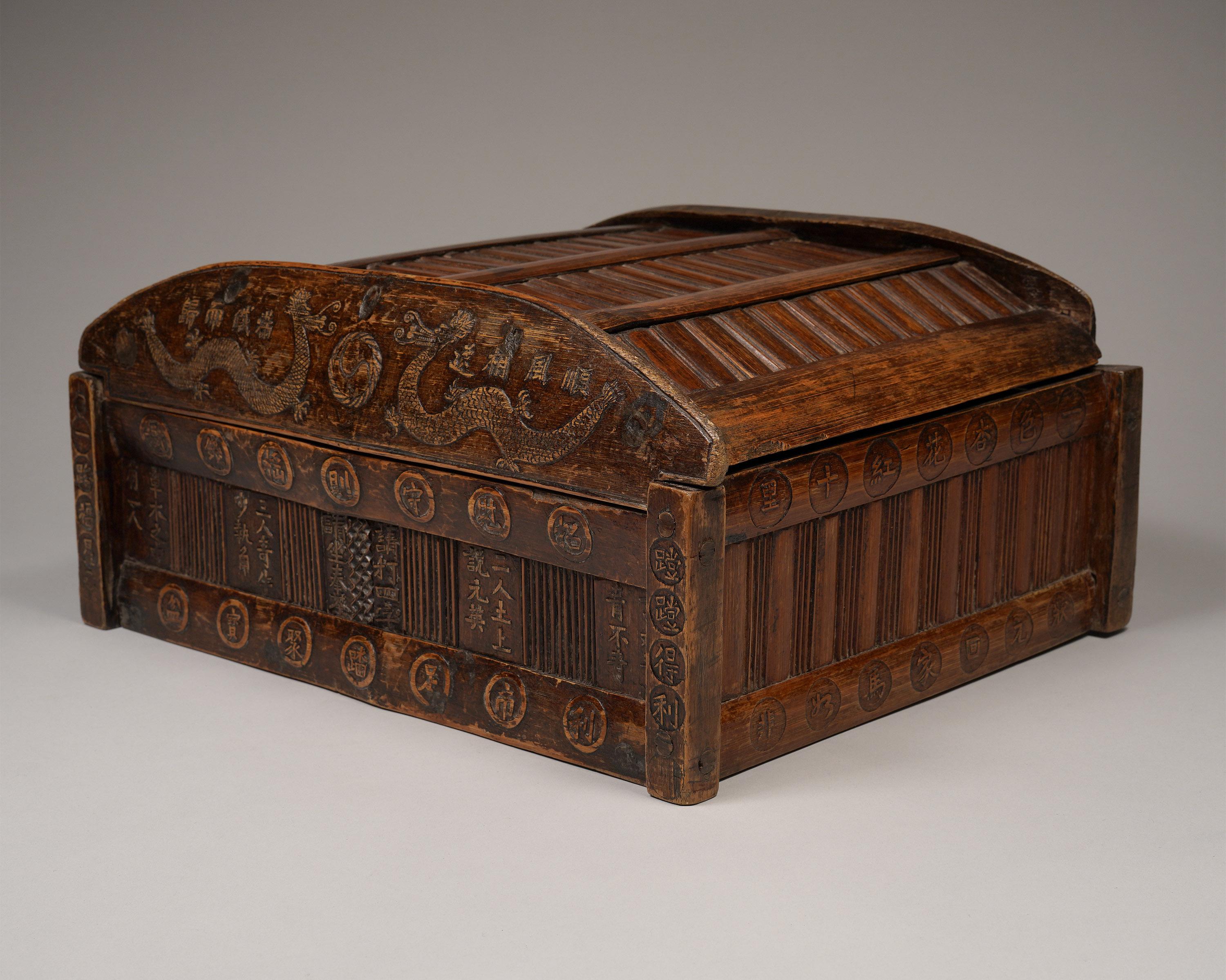 Tirelire du commerçant avec bénédictions
Chine du Sud
Fin du XIXe/début du XXe siècle
Bois, bambou
H : 7 in x W : 14.75 in x D : 11.75 in

Deux dragons entourent une perle de sagesse sur la couverture de cette boîte en bambou unique. Le texte de la