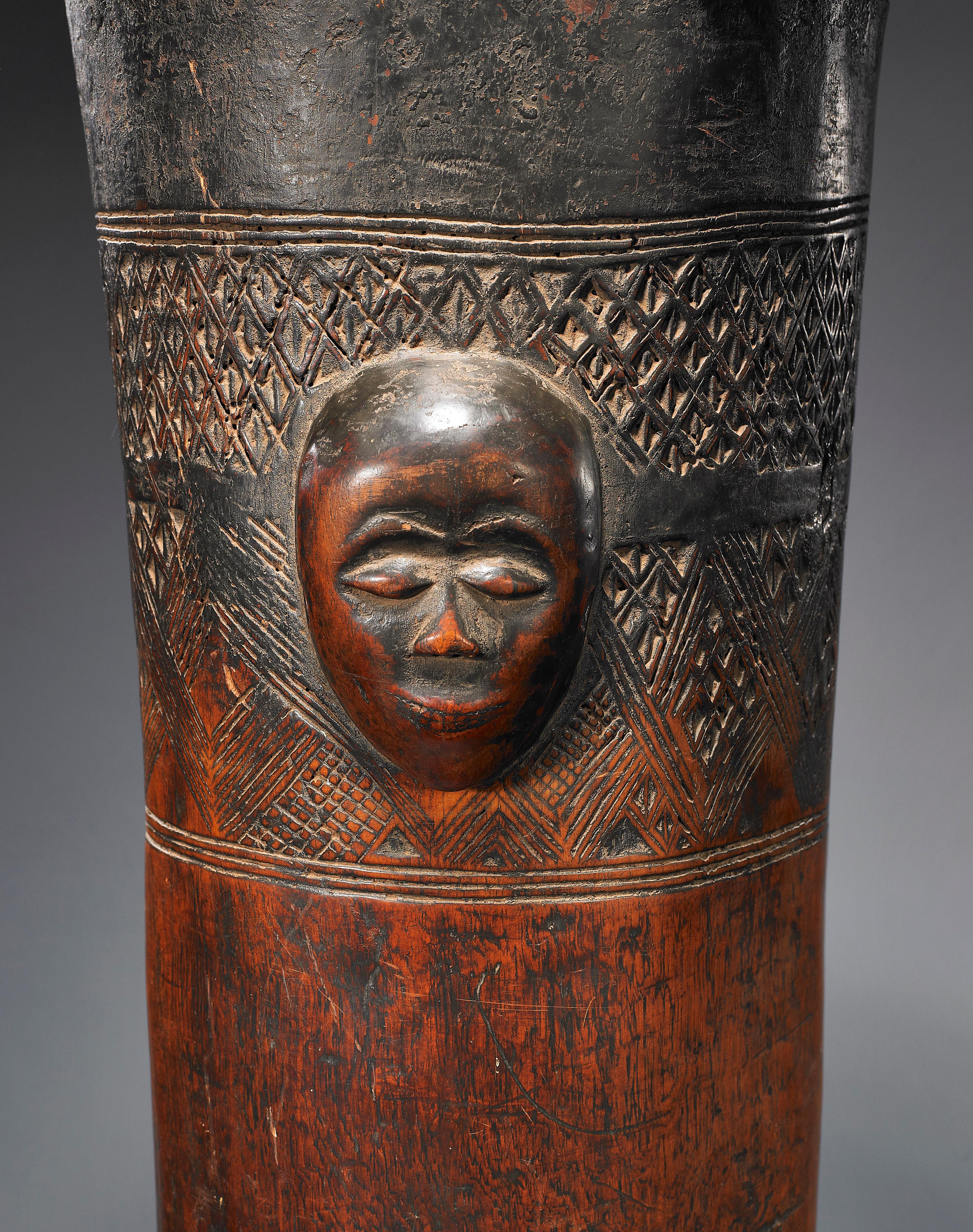 A tall and elegant wood pedestal drum with a fully-carved face on the side, and two registers of incised linear designs above and below.  It has an absolutely gorgeous honey-brown patina, that could only have come from decades of handling and