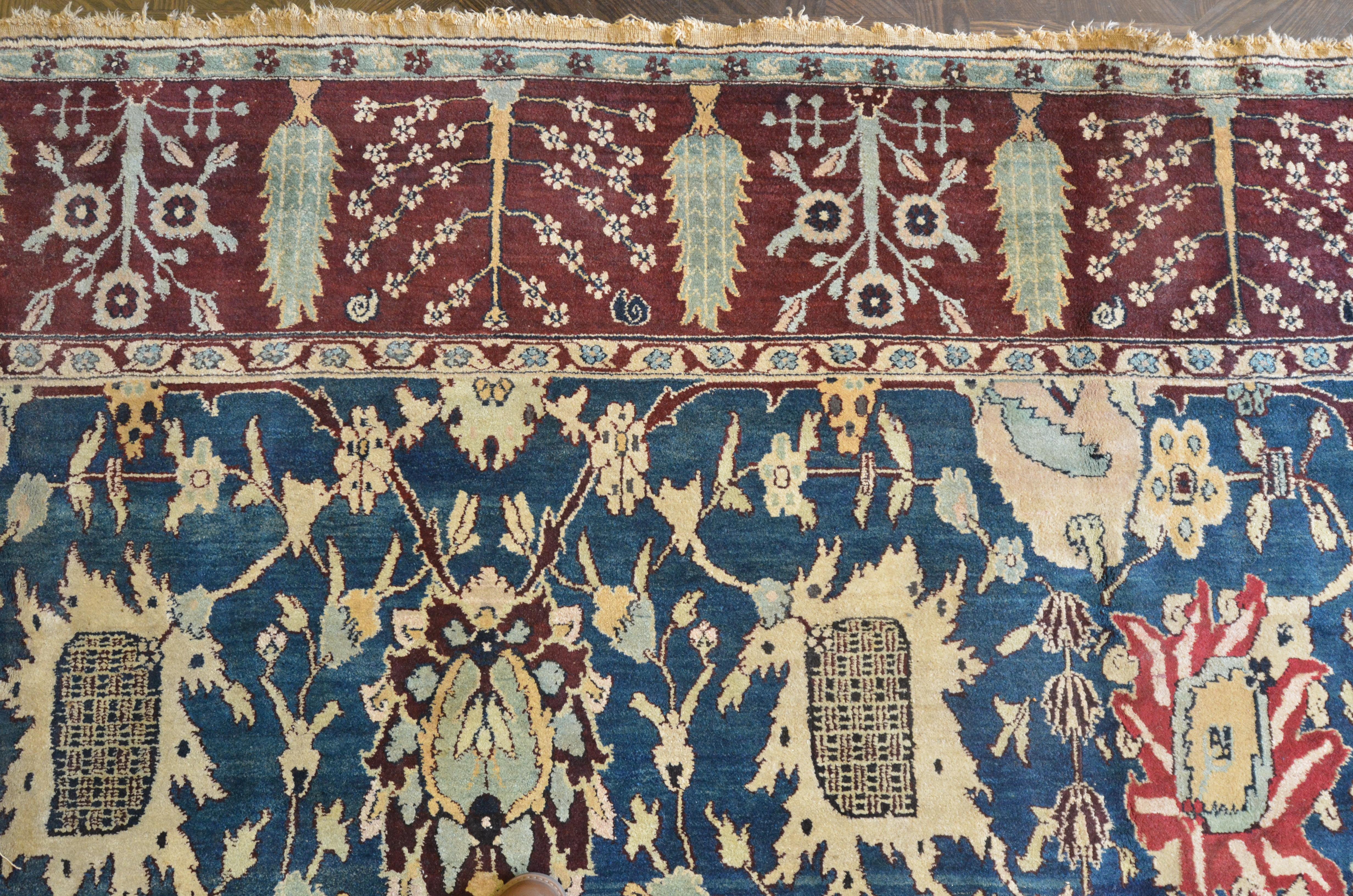 Ce tapis traditionnel indien tissé à la main à Agra présente un champ extrêmement fin de couleur bleu moyen avec un motif contrasté de palmettes et de vignes florales, dans une bordure rouge bourgogne avec des cyprès alternant avec des gerbes