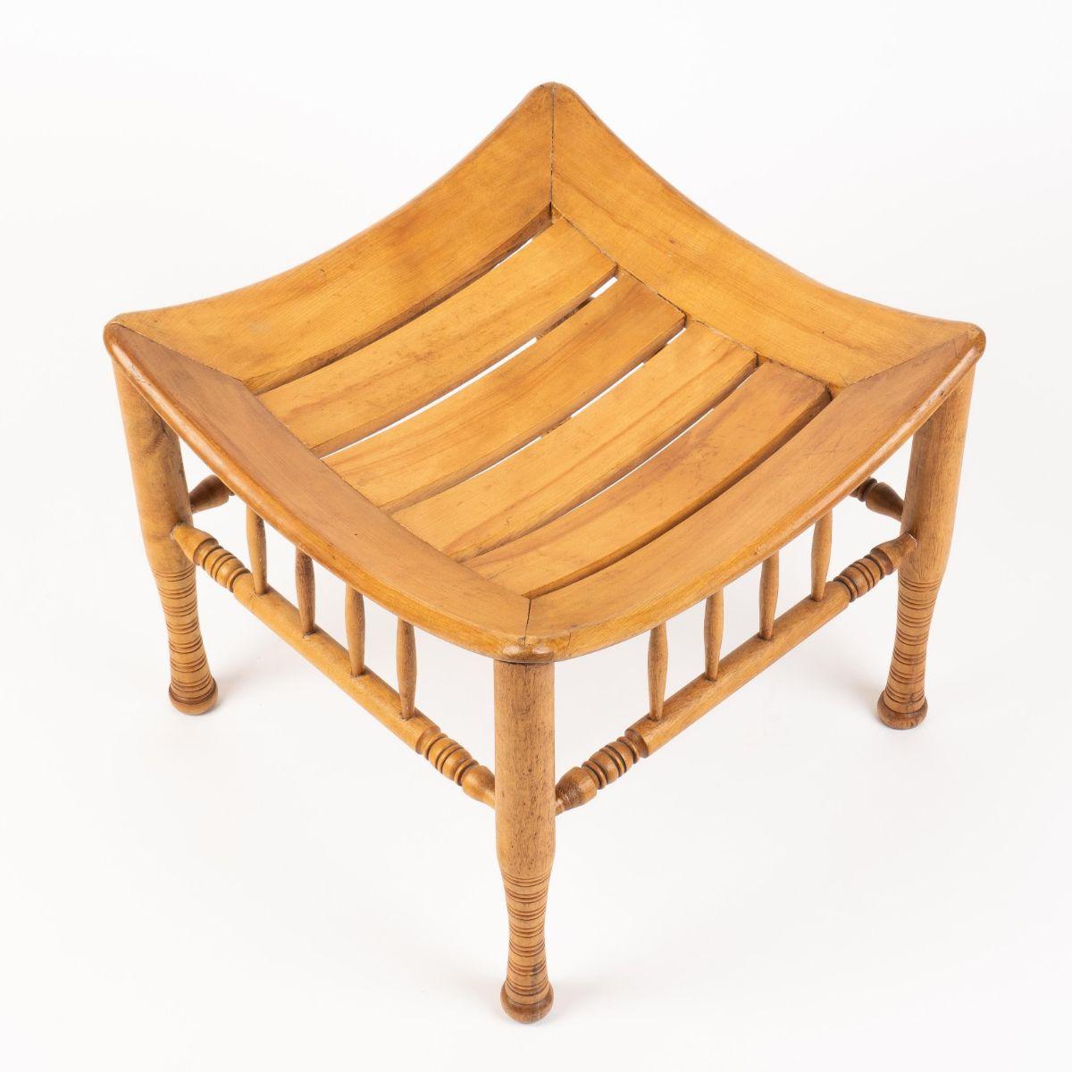 Egyptian Revival “Thebes” style stool in naturally finished birch wood, after an archeological model. The four turned legs are joined by a medial boxed stretcher each with three turned vertical braces to the cupped, frame, and slated