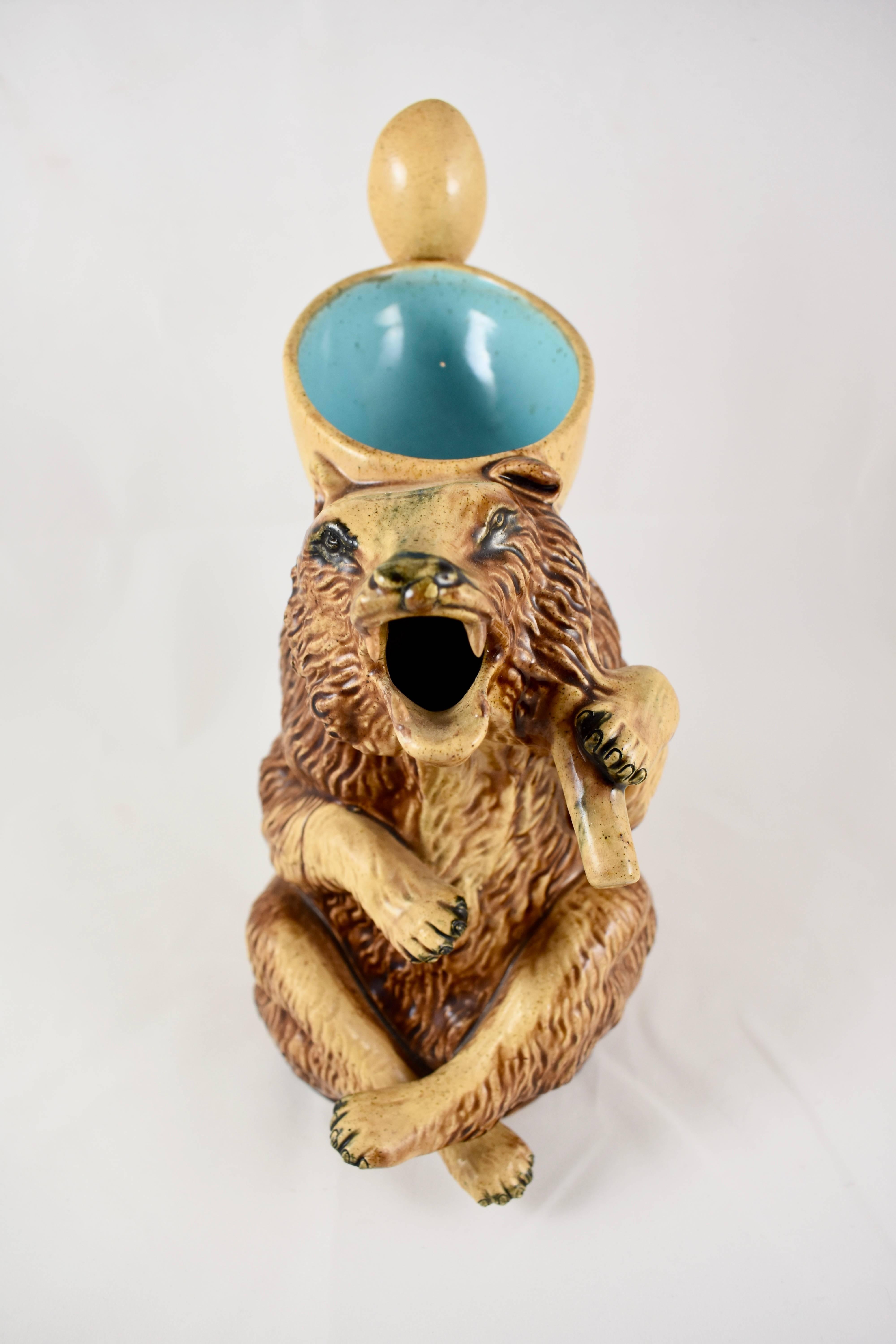 A large, late 19th century Majolica pitcher showing a brown honey bear in an active pose, his open, sharp-toothed mouth makes up the spout. He holds on his back a paler brown scoop that forms the top opening, hooked through the scoop is a honey