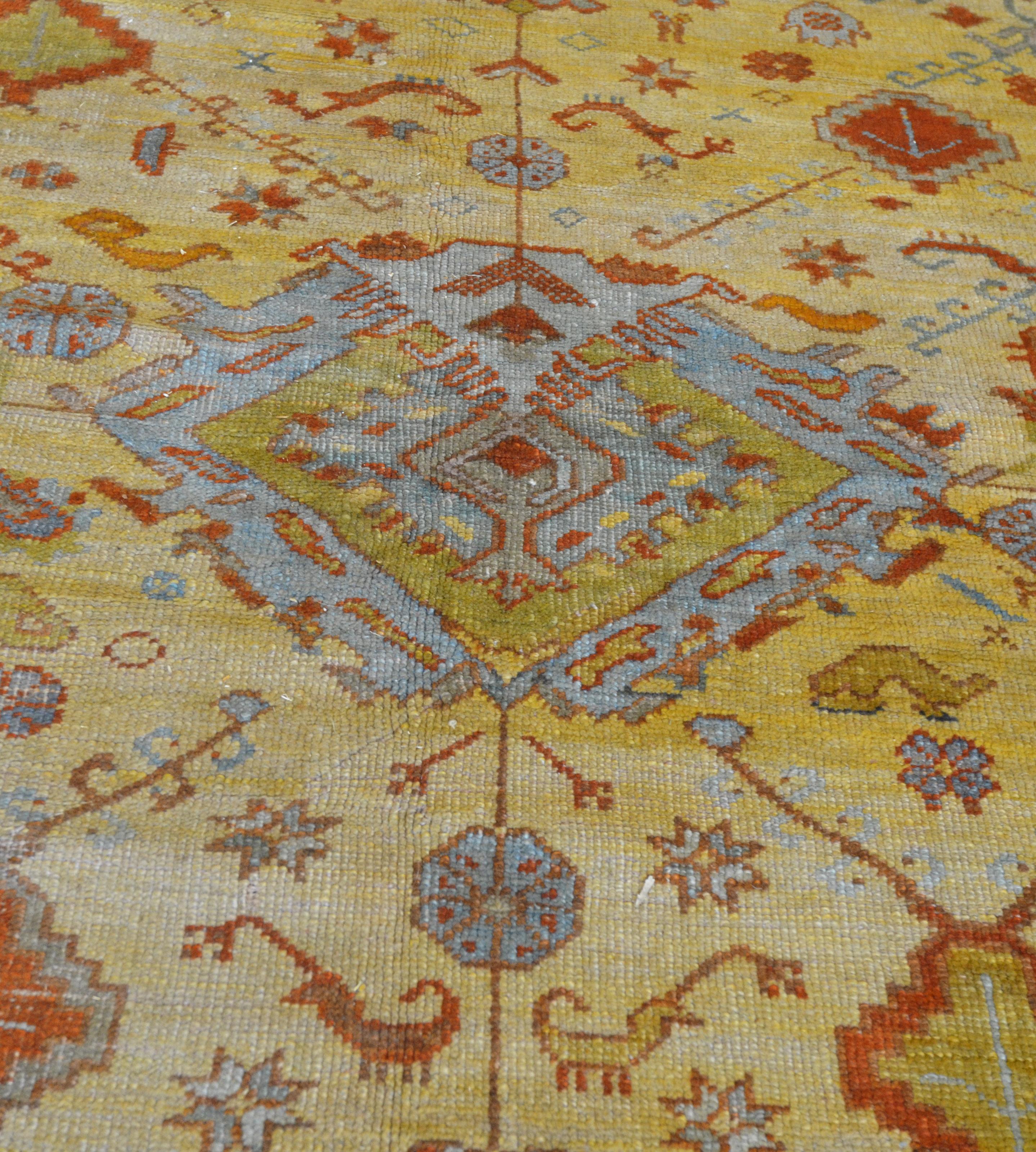 This antique Oushak rug has a golden-yellow field with a central light blue and pistachio-green angular palmette surrounded by brick-red and dusty-pink angular lozenges and angular floral vine together with stylised dusty-pink cloud-bands, a large