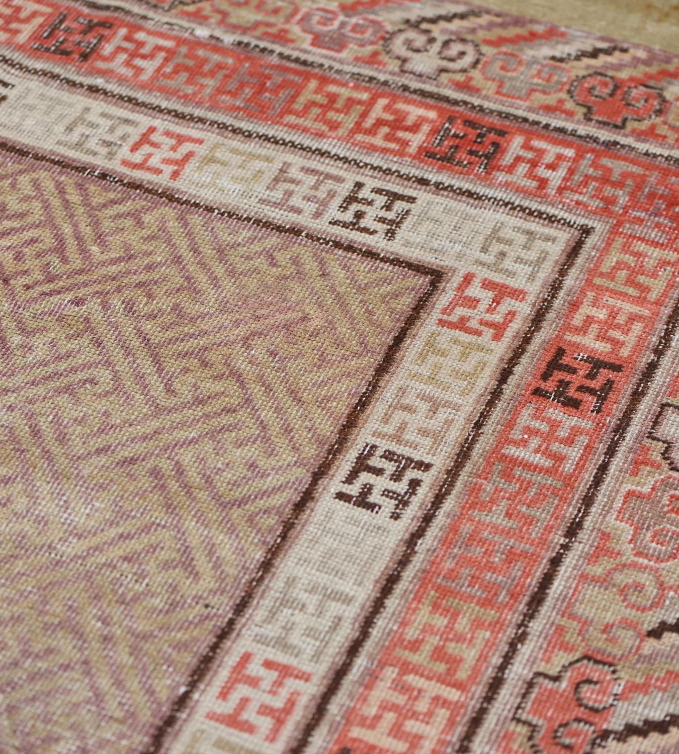 This antique Khotan rug has a light green field with an overall design of aubergine-purple interlocking lattice-weave, enclosed by a double band of ivory and pale red stripes with chocolate-brown, aubergine-purple and soft grey T-shaped lozenges