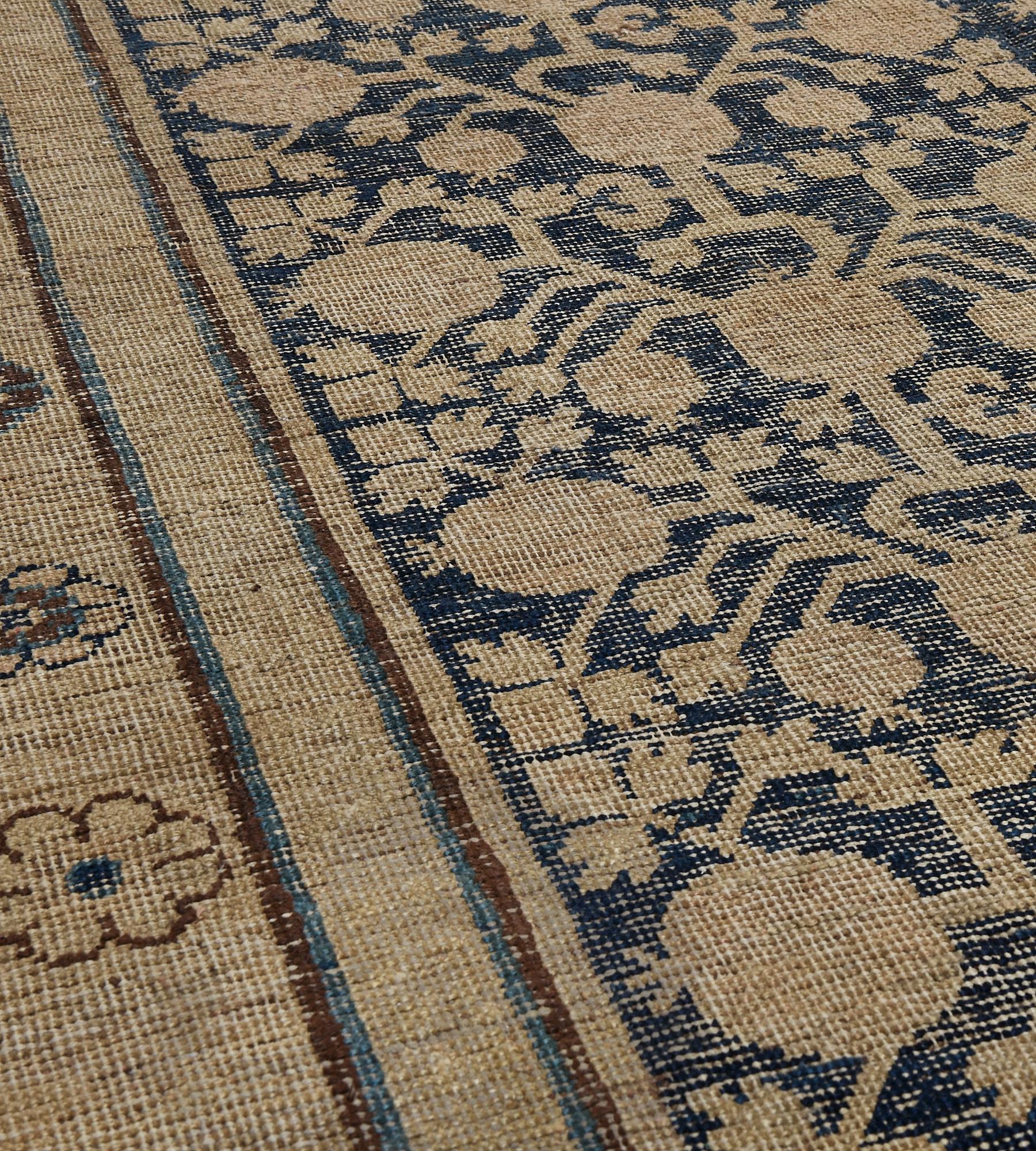 This antique Khotan rug has a deep indigo-blue field with an overall sandy-brown pomegranate and floral vine, in an outer broad sandy-brown border with cloud-motifs alternating with a plant motif, inner key-pattern, flowerhead and plain stripes.