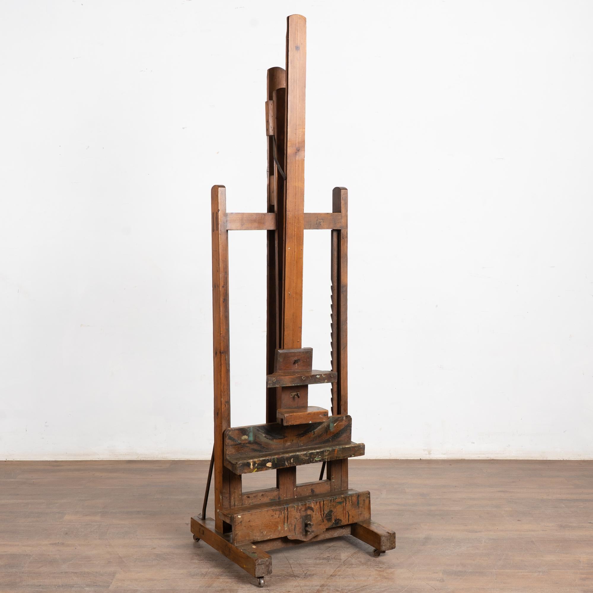 Questo cavalletto per artisti vintage poggia su quattro rotelle. Rivela anni di utilizzo, come testimoniano le numerose macchie di vernice, i graffi, le ammaccature e le aree di sofferenza e usura.
Tutte le aree di sofferenza sono dovute all'età e