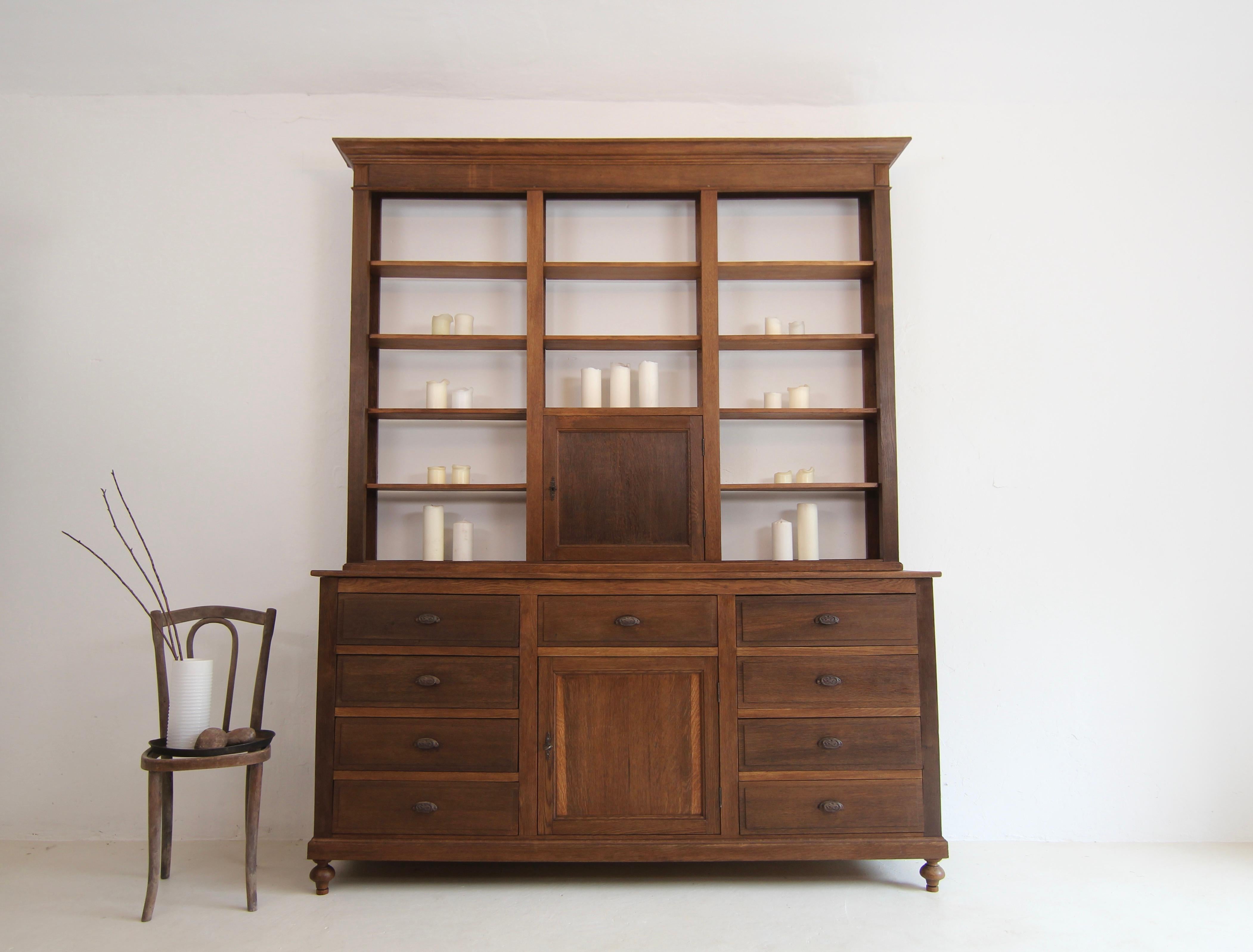 A authentic German or Belgian apothecary's shop cabinet from around 1900.

Two-part side-moulded oak body consisting of base and shelf unit.

The base cabinet has a door and 9 drawers under a slightly protruding top panel. The flat open shelf with